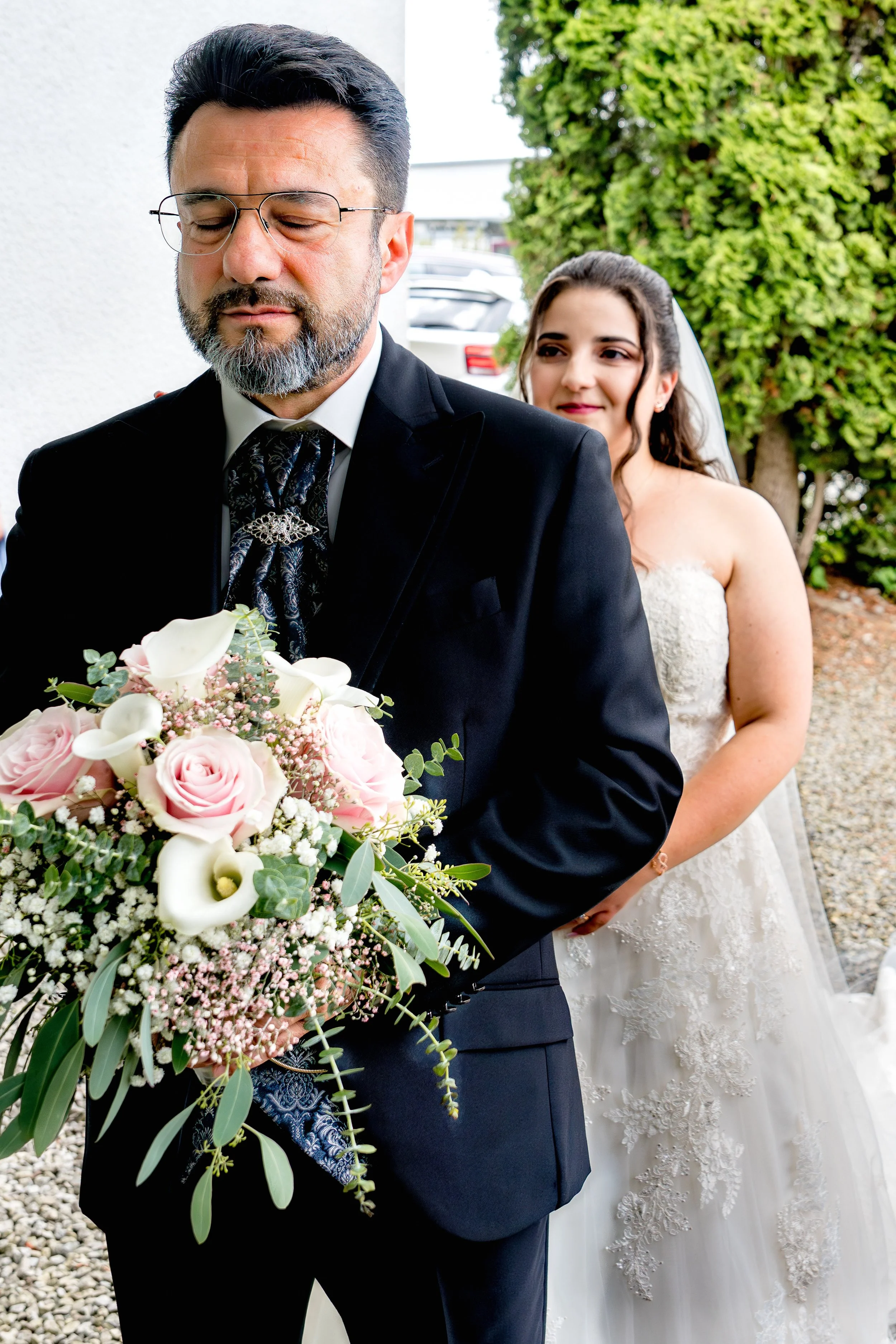 Die Braut steht hinter ihrem Vater und ist bereit für den First Look. Der Vater hat die Augen fest verschlossen und ist sichtlich aufgeregt. Er hält den Brautstrauß in seiner Hand. 