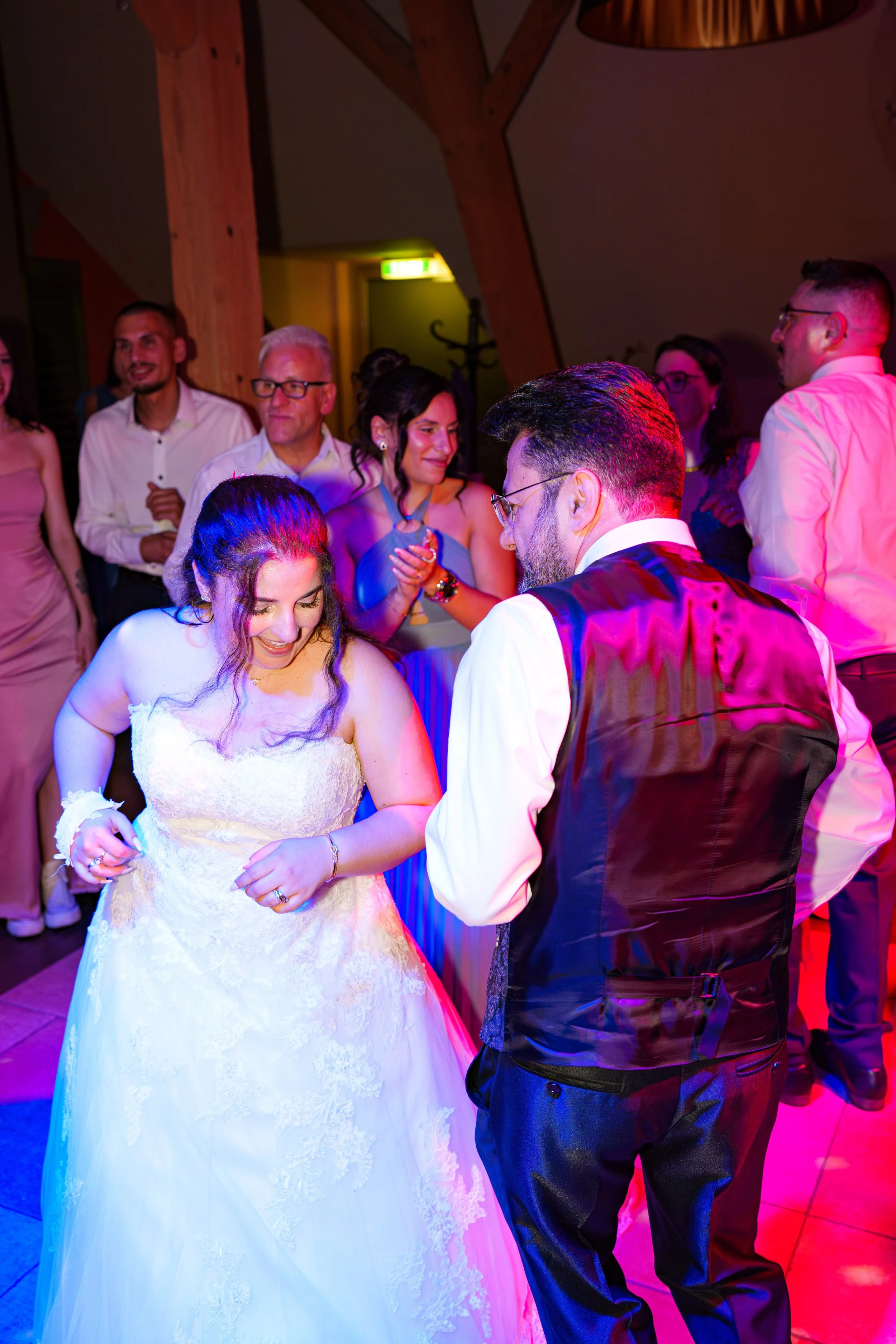 Braut und Bräutigam tanzen auf der Hochzeitsfeier mit lachenden Gästen im Hintergrund, farbig beleuchteter Raum.
