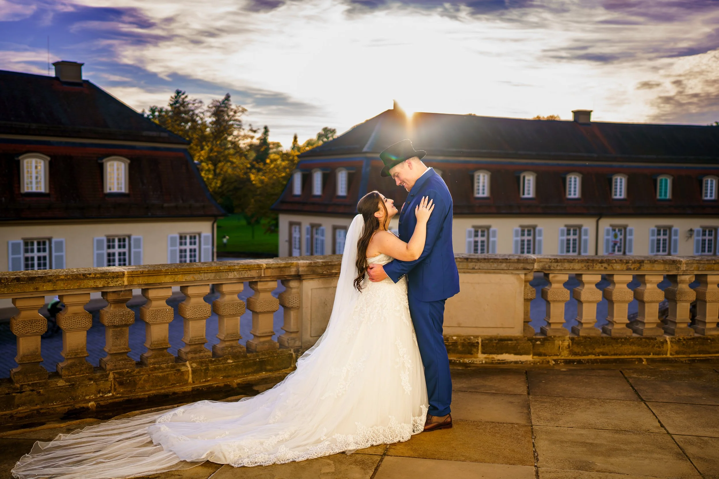 Ein Brautpaar steht auf einer Terrasse bei Sonnenuntergang, umarmt sich, im Hintergrund alte Gebäude und Bäume.