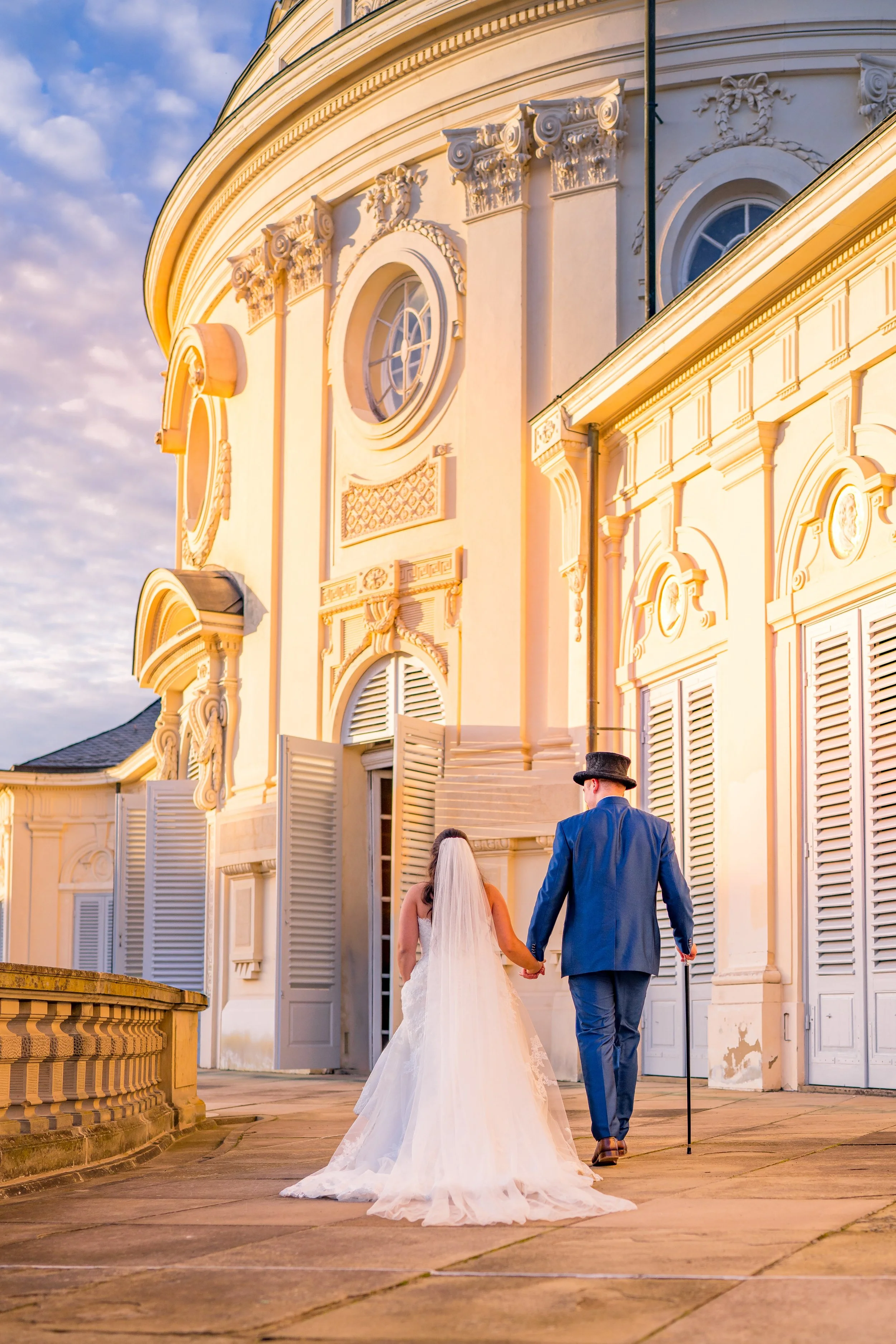 Ein Brautpaar beim Spaziergang vor einem hell erleuchteten barocken Gebäude bei Sonnenuntergang, die Frau trägt ein weißes Brautkleid mit Schleier, der Mann trägt einen blauen Anzug und einen Hut.