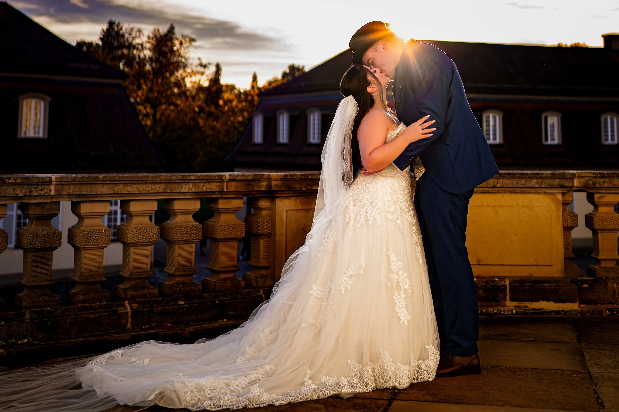 Ein Brautpaar küsst sich bei Sonnenuntergang auf einer Terrasse, Braut trägt ein weißes Hochzeitskleid mit langer Schleppe, Bräutigam trägt einen dunklen Anzug, im Hintergrund Dach und Baumkronen.