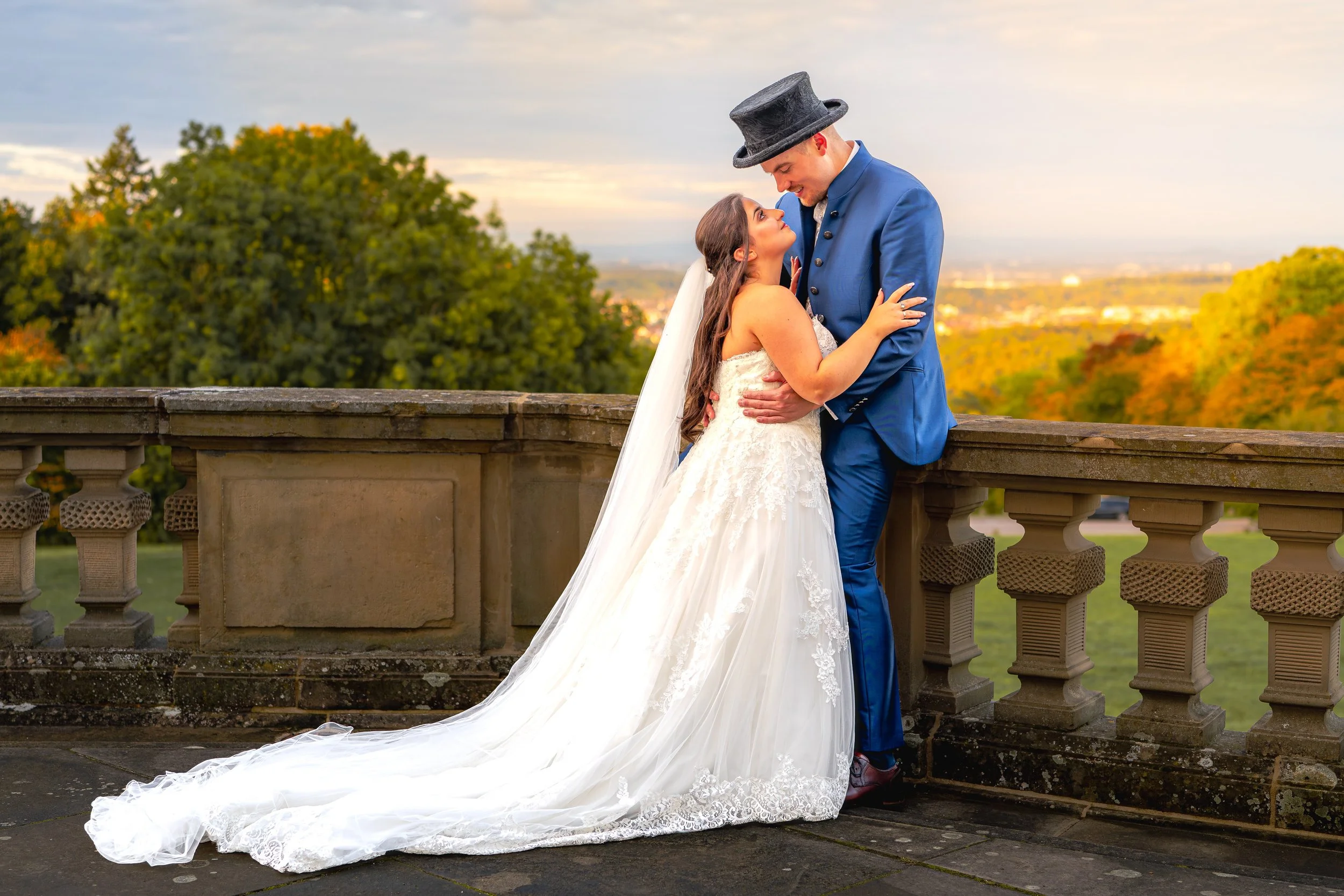 Ein verheiratetes Paar in Hochzeitskleidung steht sich auf einer Brüstung gegenüber, der Himmel ist leicht bewölkt und die Bäume im Hintergrund zeigen Herbstfarben.
