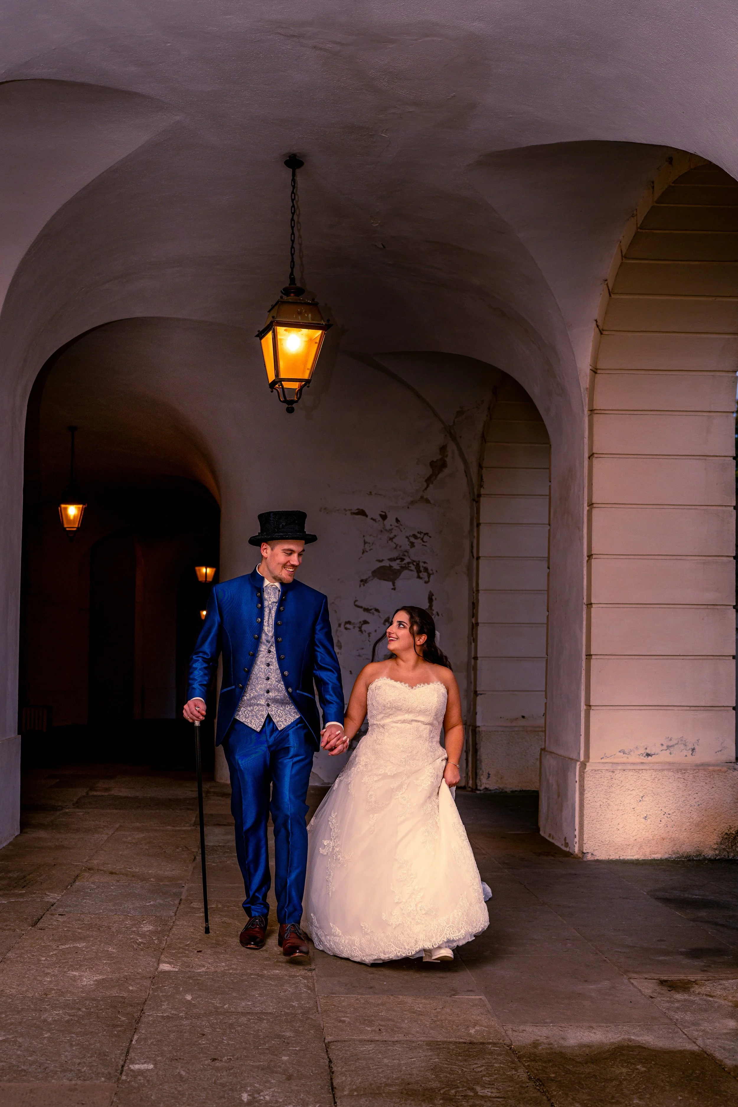 Ein Paar in Hochzeitskleidung, ein Mann in einem blauen Anzug und eine Frau in einem weißen Brautkleid, spazieren Hand in Hand unter einer alten Arkade, beleuchtet von Laternen.