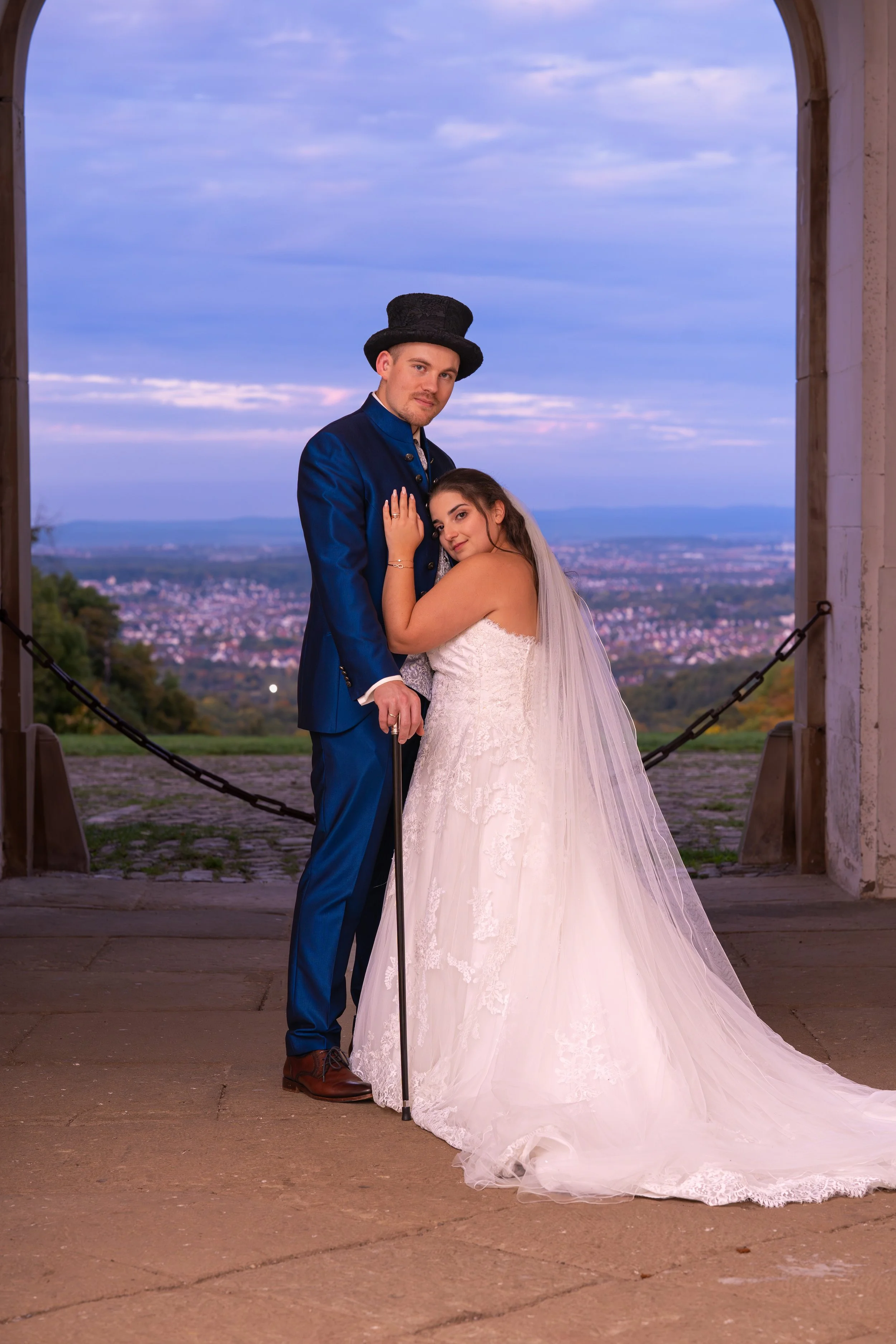 Ein Brautpaar steht vor einer Kulisse mit Blick auf eine Stadt bei Sonnenuntergang. Der Bräutigam trägt einen blauen Anzug und einen Zylinder, die Braut trägt ein weißes Brautkleid mit langem Schleier.