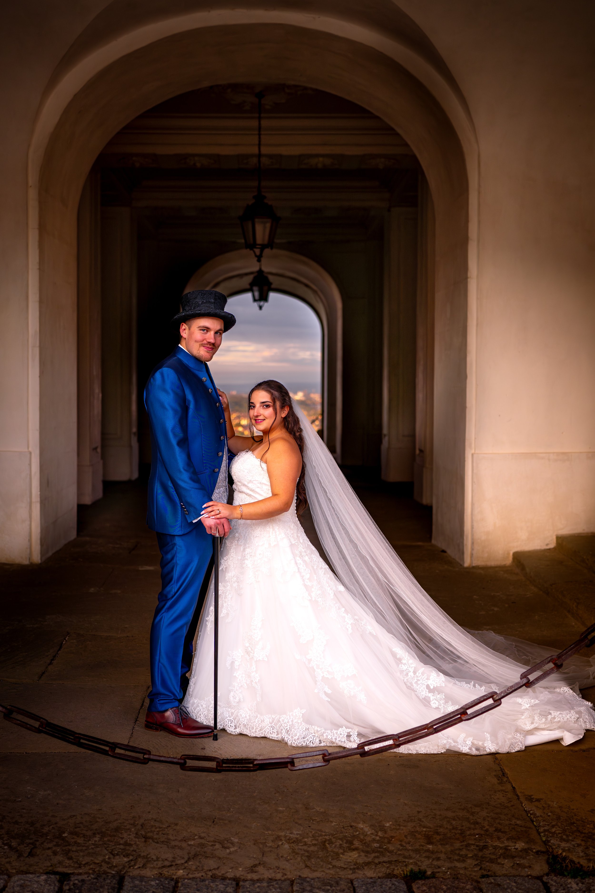 Ein frisch vermähltes Hochzeitspaar posiert unter einer Arkade mit Laternen, im Hintergrund eine Stadt bei Sonnenuntergang.