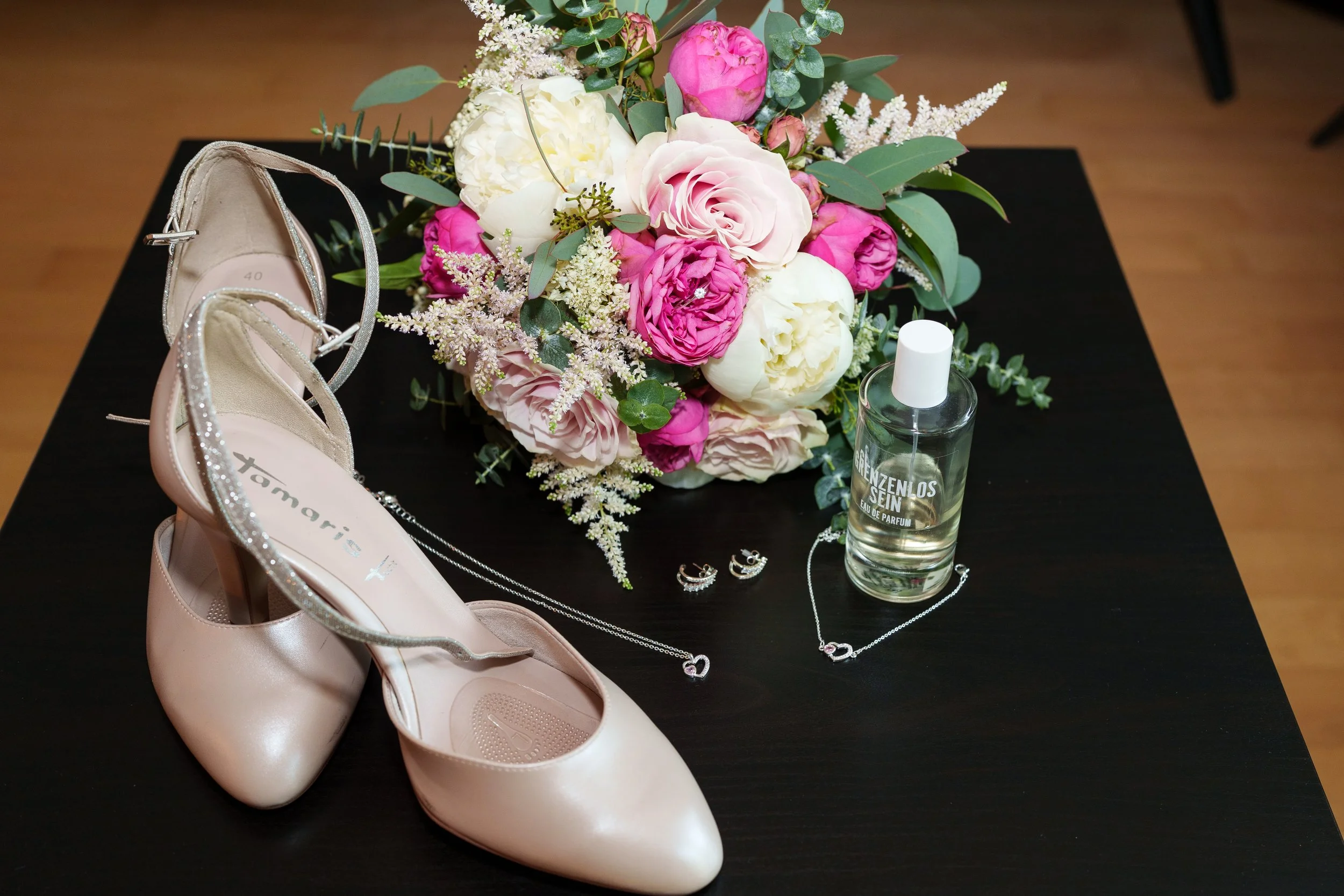 Kleider, Schuhe, Schmuck, Parfüm und ein Blumenstrauß auf einem schwarzen Tisch, wahrscheinlich für eine Hochzeit vorbereitet.
