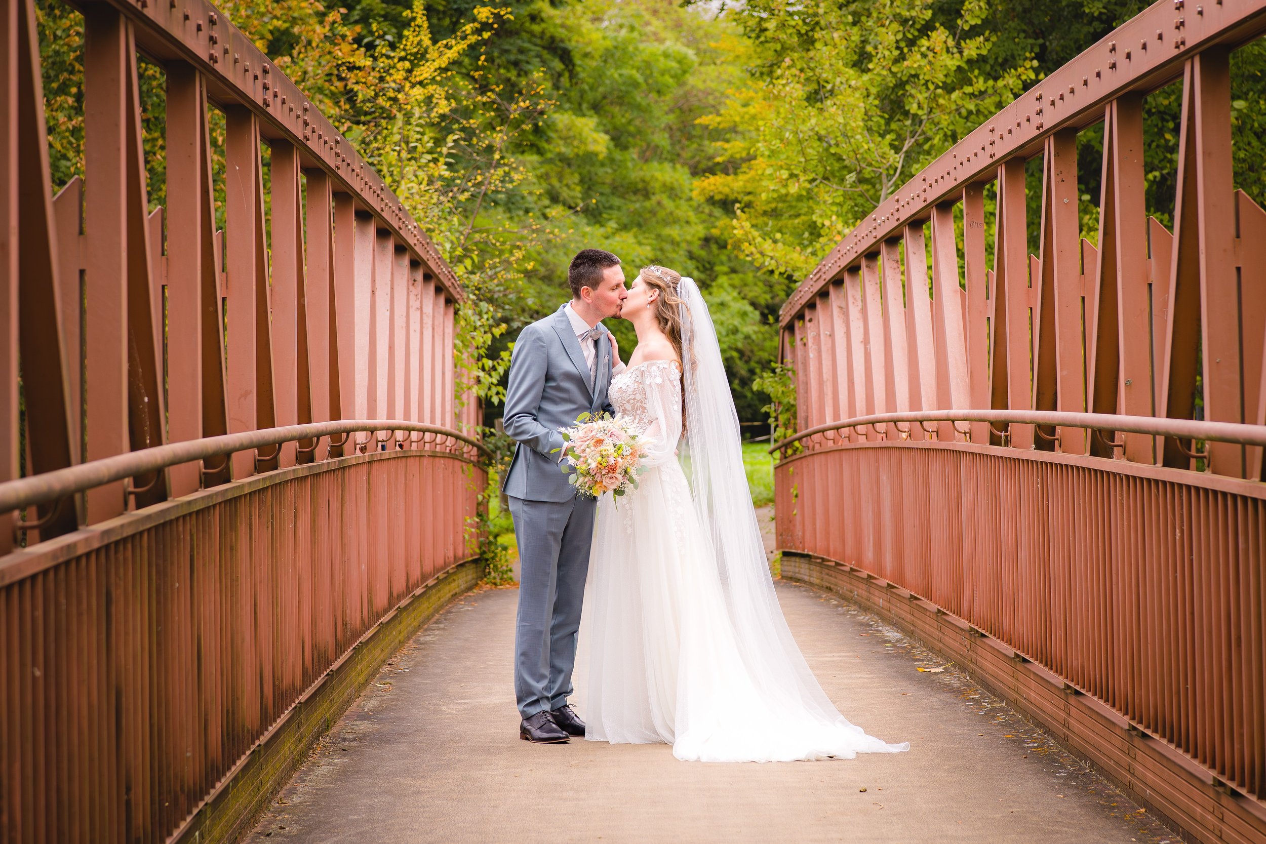 Ein Brautpaar im Hochzeit-Outfit, das sich auf einer Brücke mit roten Geländern küsst, umgeben von grünen Bäumen bei sonnigem Wetter.