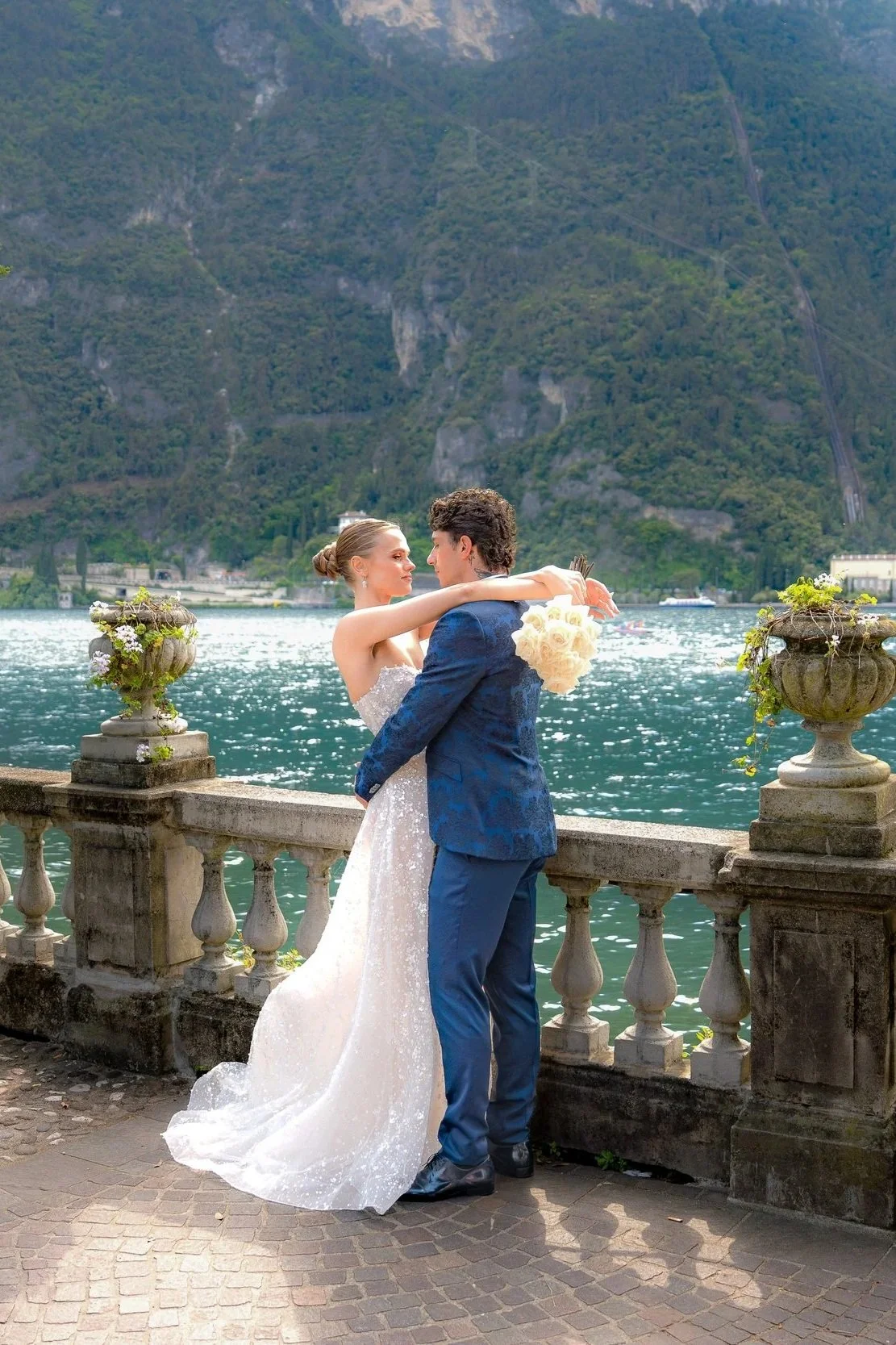 Ein Brautpaar umarmt sich am Ufer eines Sees mit Bergen im Hintergrund, die an einem sonnigen Tag festliche Kleidung tragen.