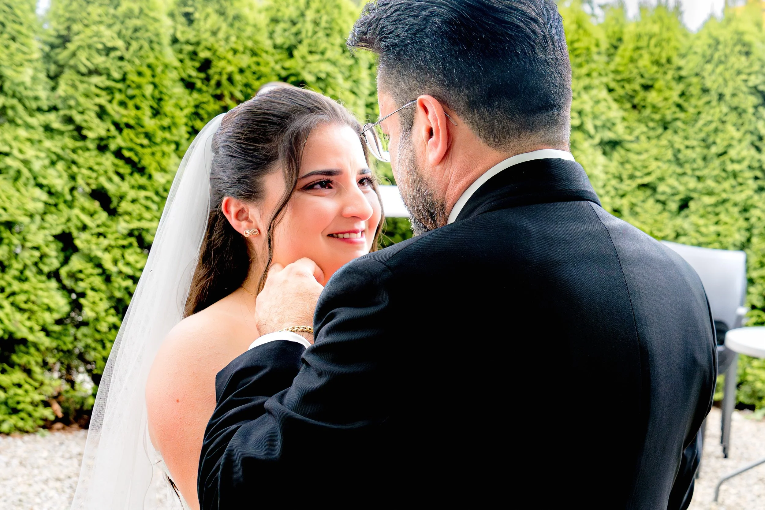 Der Vater sieht das erste mal seine Tochter im Brautkleid. Die Braut weint und der Papa streichelt ihr liebevoll über die Wange. Im Hintergrund sind grüne Bäume sichtbar.
