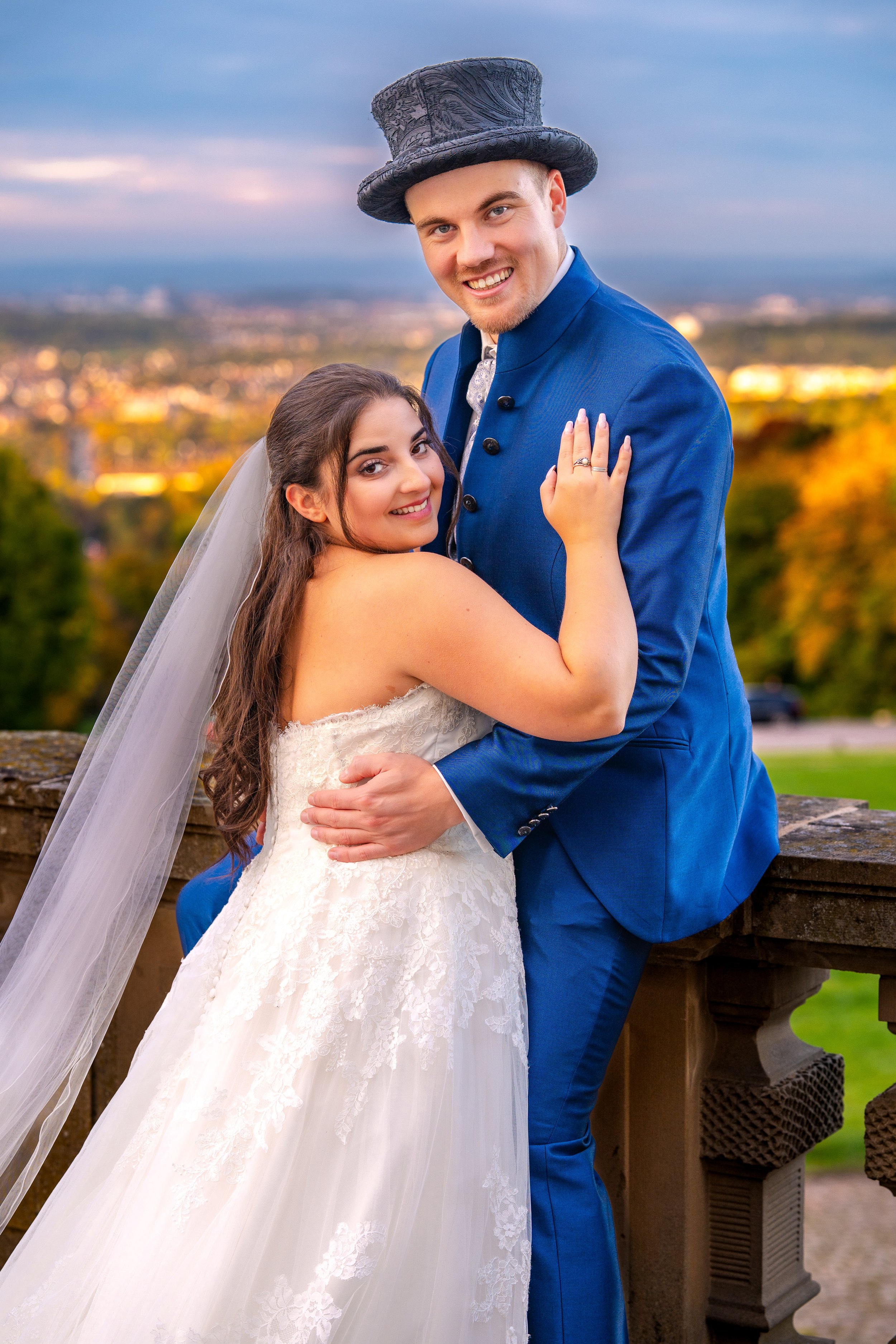 Ein glückliches Brautpaar steht auf einer Terrasse mit Blick auf die Stadt im Hintergrund, der Mann trägt einen blauen Anzug mit einem schwarzen Hut, die Frau trägt ein weißes Hochzeitskleid mit Schleier.