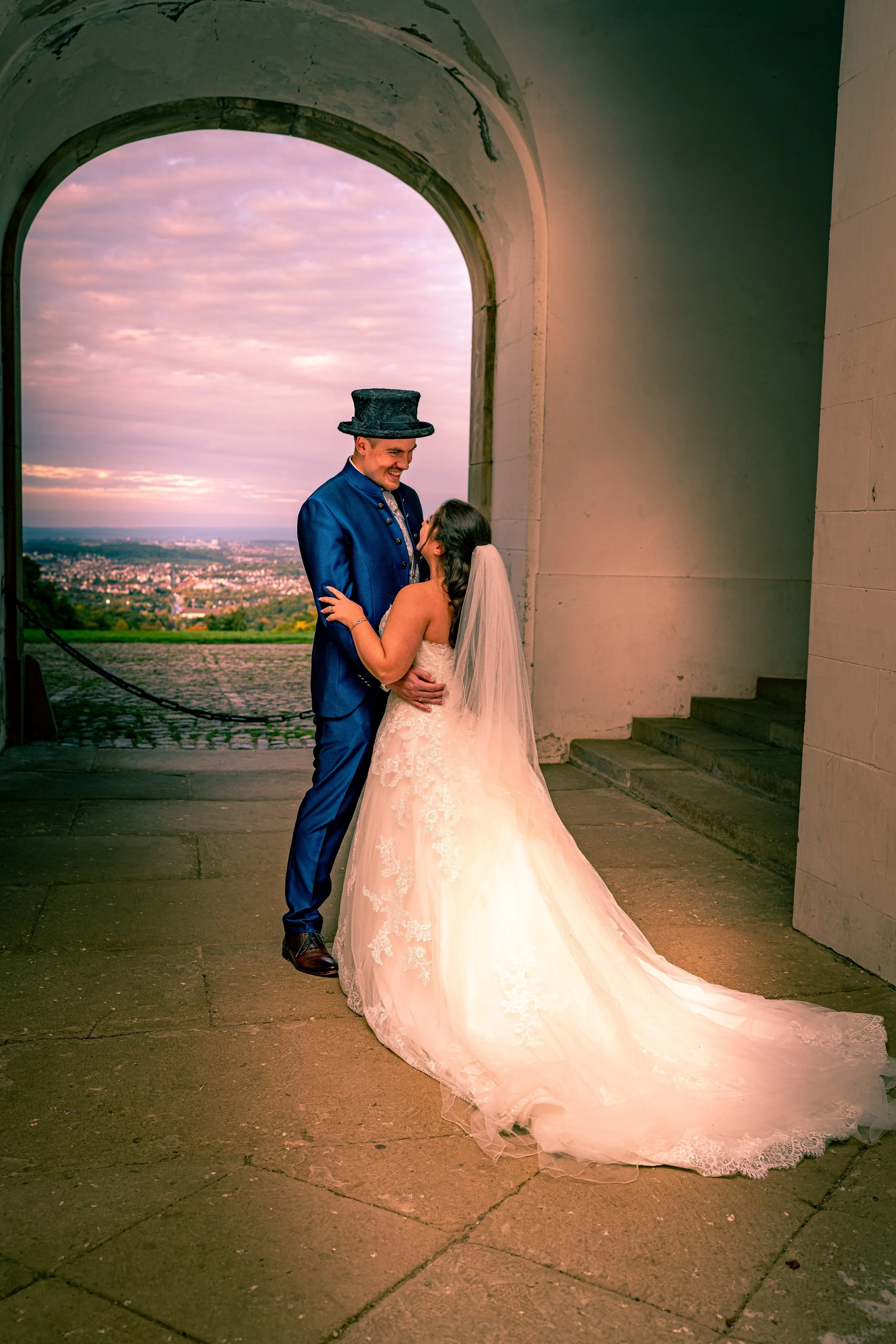 Ein Brautpaar bei einer Hochzeit, der Bräutigam trägt einen blauen Anzug und einen Zylinder, die Braut trägt ein weißes Hochzeitskleid und einen Schleier, sie stehen sich gegenüber unter einem großen Torbogen vor einer malerischen Landschaft bei Sonn