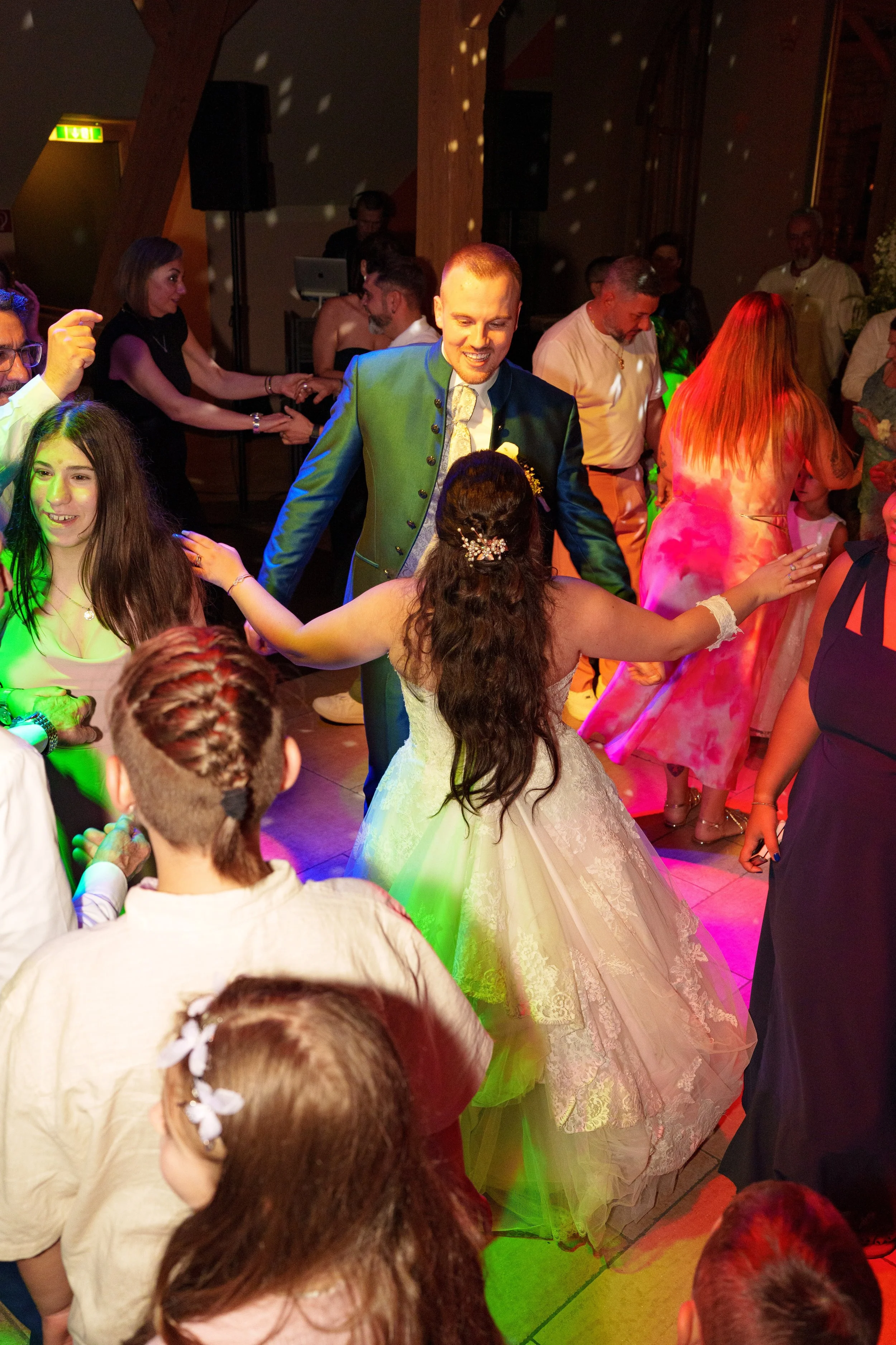 Ein Mann in einem grünen Anzug tanzt mit einer Braut in einem weißen Hochzeitskleid auf einer Party, umgeben von anderen tanzenden Gästen in einem festlich beleuchteten Raum.