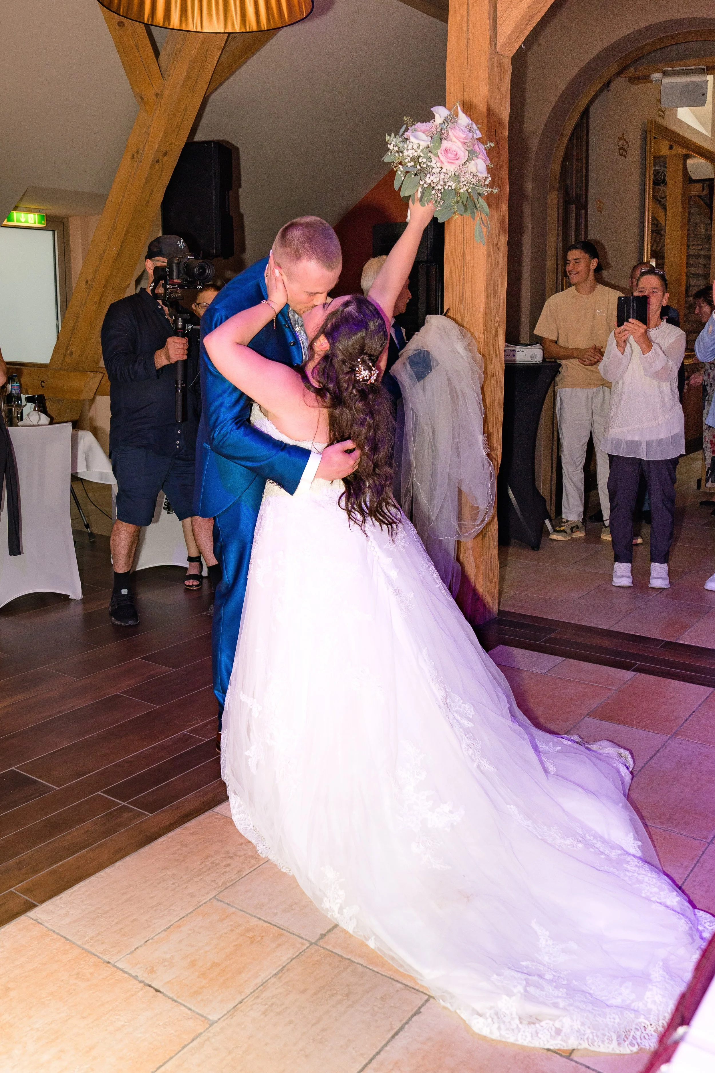 Ein Brautpaar, dass im Saal der Hofgut Domäne umgeben von ihren Gästen sich im arm hält und küsst. Sie hält den Brautstrauß nach oben. 