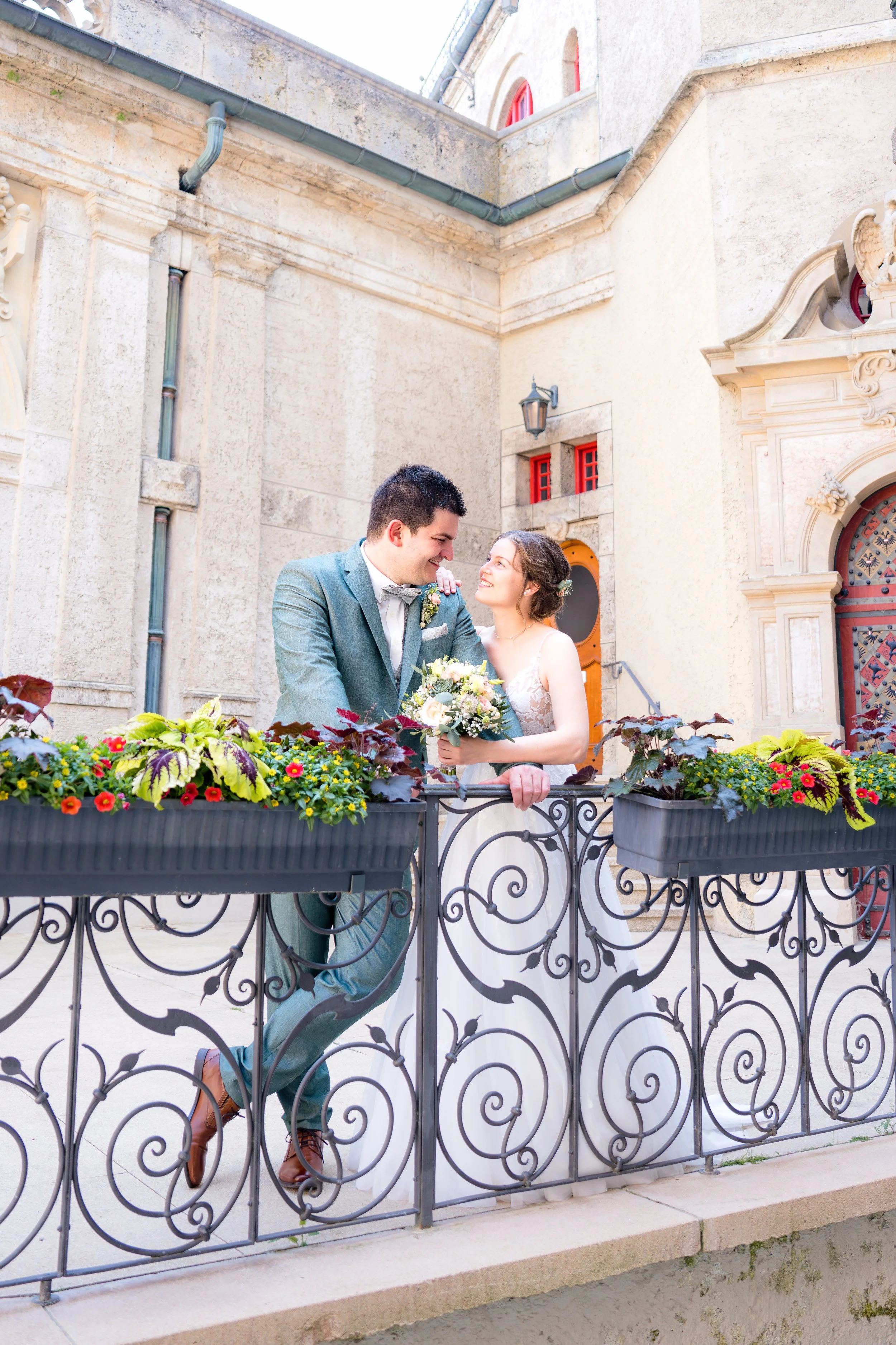 Ein glückliches Brautpaar steht auf einem Balkon mit dekorativen Metallgeländern, umgeben von Blumen, vor einer alten, historischen Gebäude in einer Innenstadt.