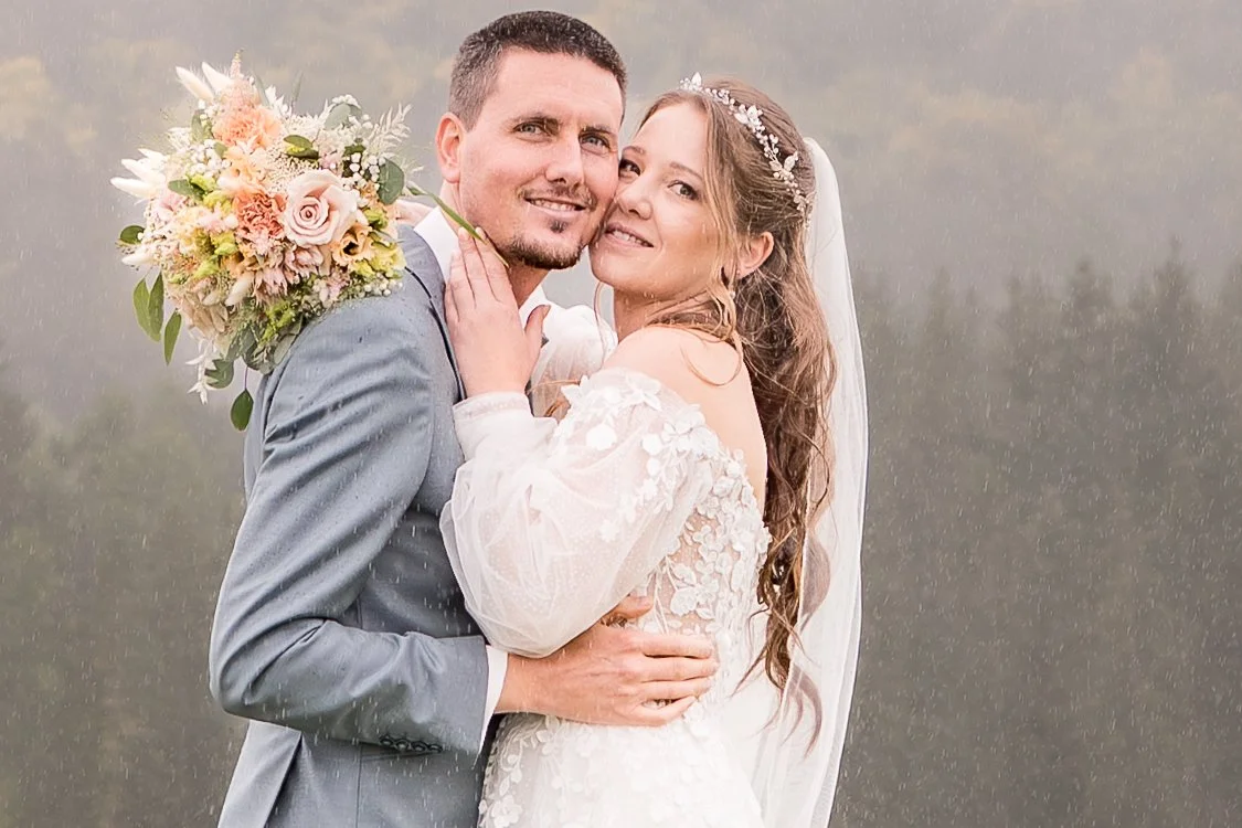 Ein lächelndes Brautpaar vor einem bewölkten Himmel, die Braut trägt ein weißes Kleid mit Spitze und eine Diadem, der Bräutigam trägt einen grauen Anzug, sie halten sich eng umarmt.