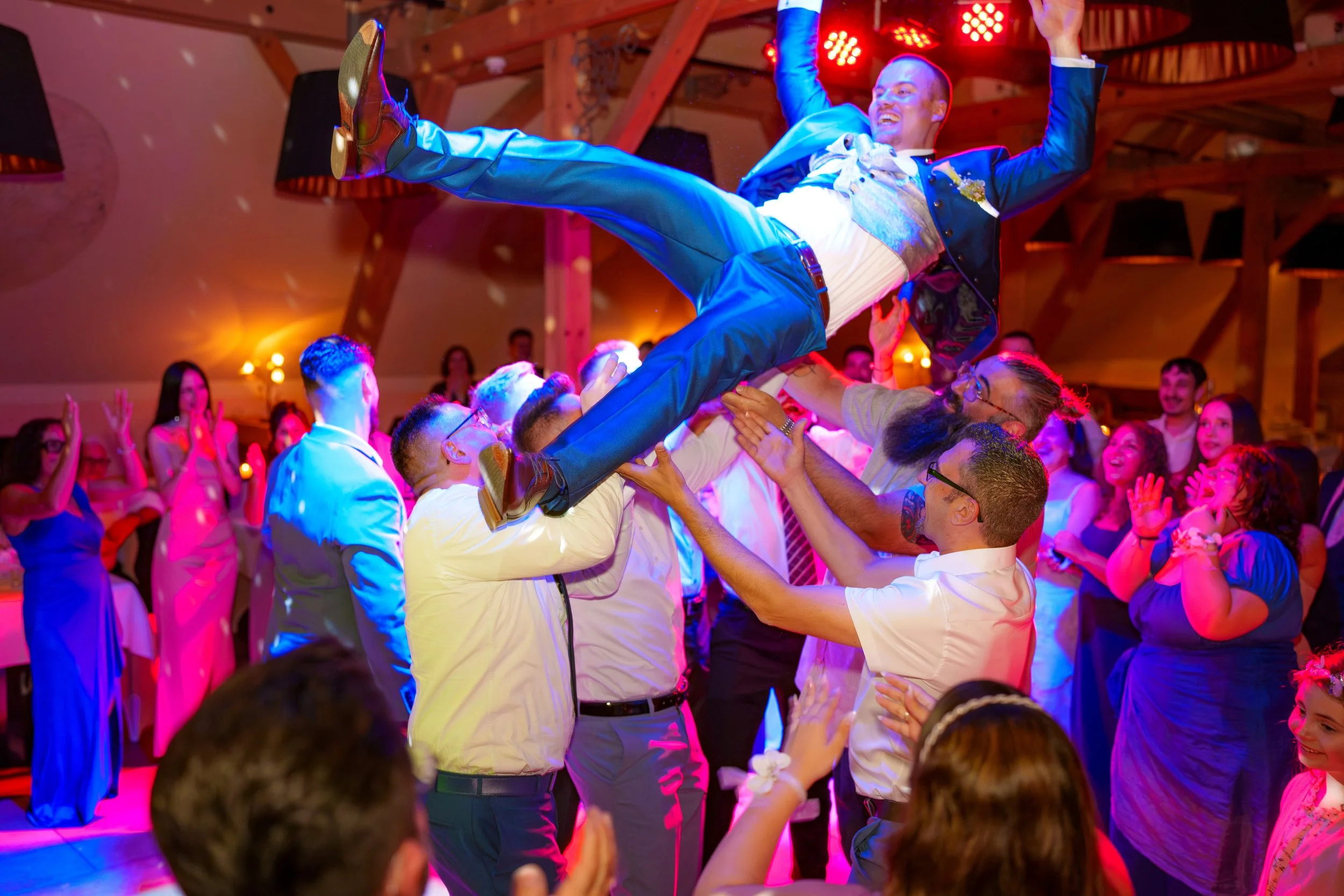 Junge Mann in blauem Anzug und weißen Hemd wird in die Luft gehoben bei einer Festlichkeit, umgeben von lachenden Menschen im festlichen Rahmen mit buntem Licht