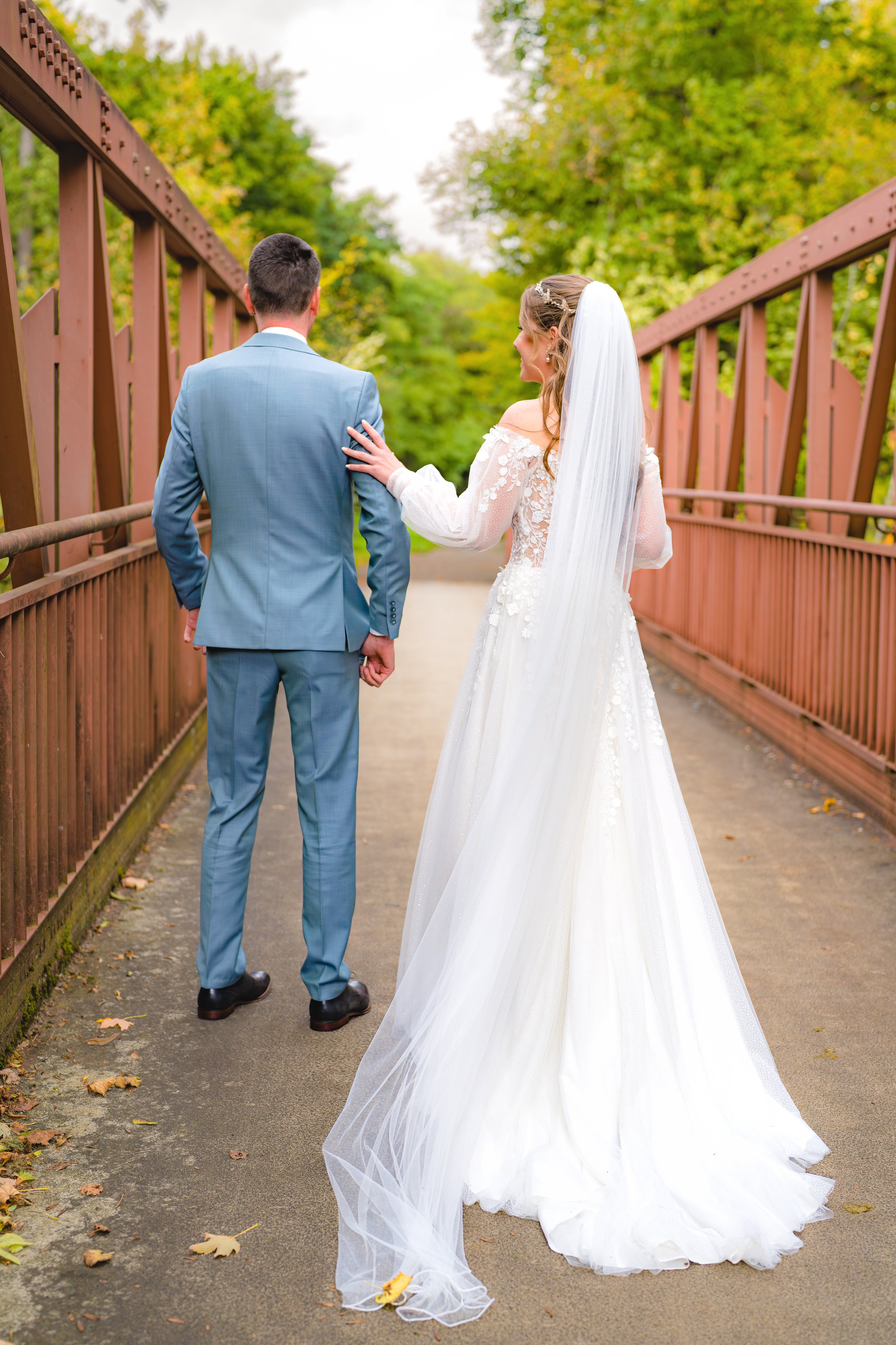 Braut und Bräutigam laufen auf einer Brücke im Park, das Brautkleid hat einen langen Schleier und die Umgebung ist grün.
