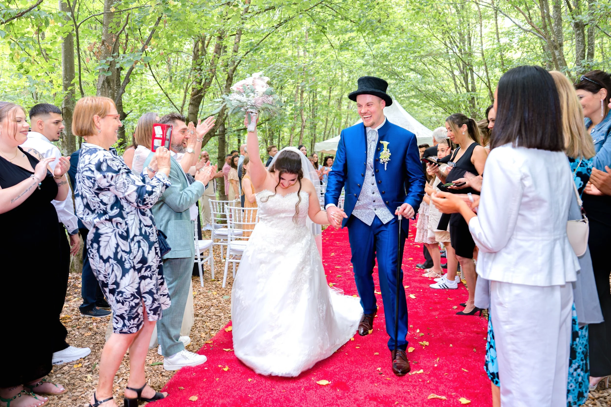 Ein Brautpaar läuft lachend zusammen auf einem roten Teppich durch einen Wald, umgeben von fröhlichen Gästen bei ihrer Hochzeit.