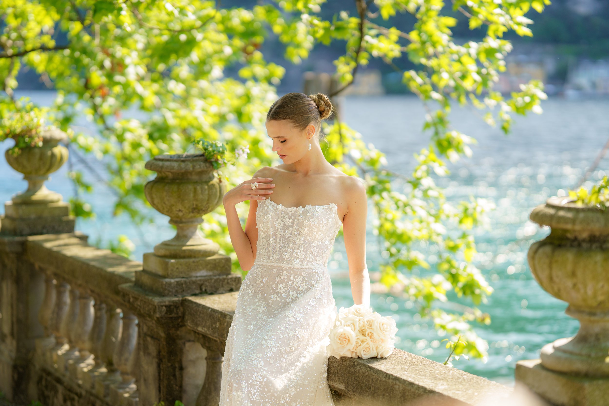 Eine brautliche Frau in einem weißen, glitzernden Brautkleid mit einem Brautstrauß aus weißen Rosen, die an einem sonnigen Tag am Wasser steht.