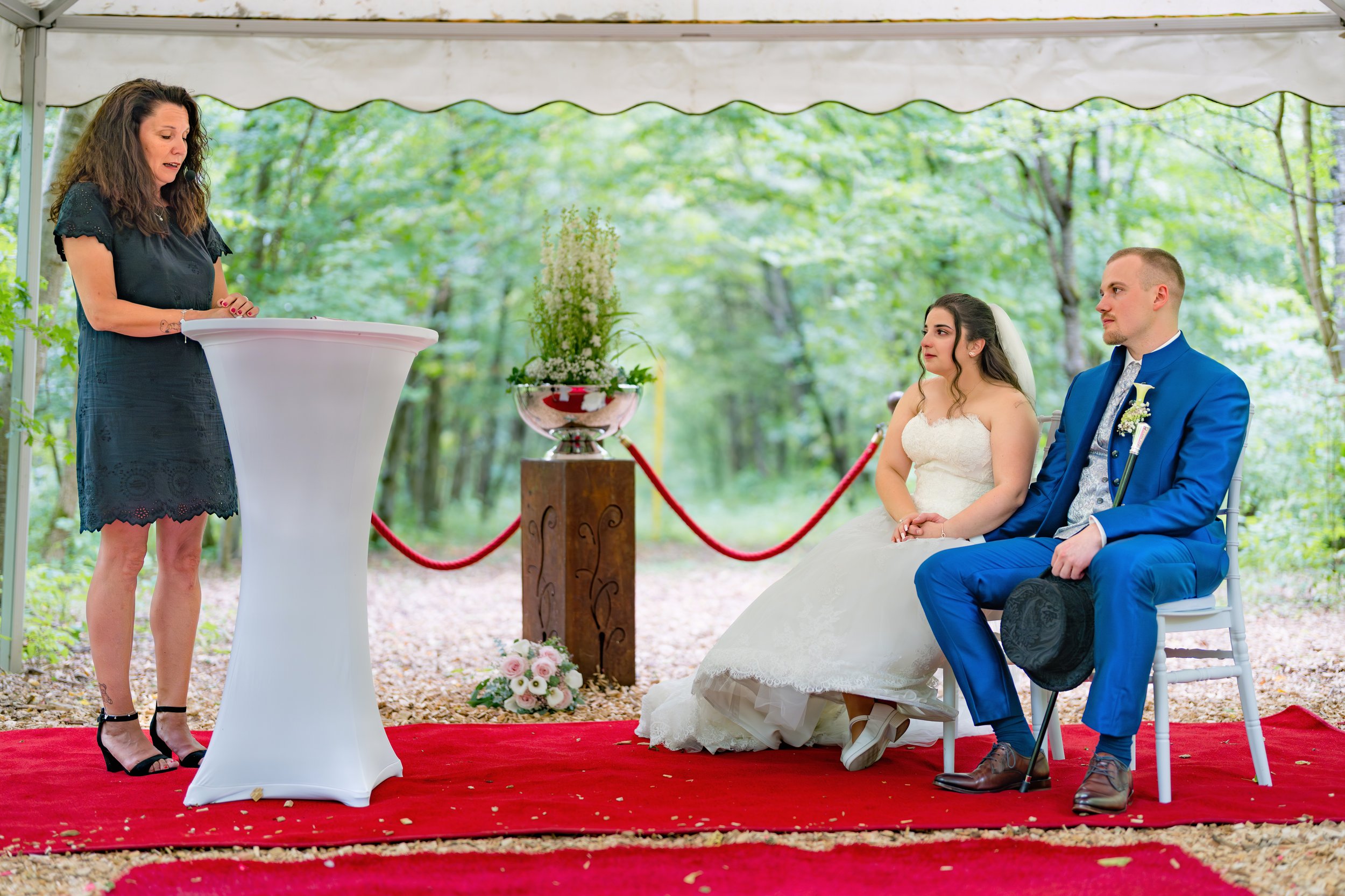 Ein Brautpaar bei einer freien Trauung im Fasanenwald in Hechingen, sitzend auf weißen Stühlen, mit einer freien Traurednerin, die spricht. Das Brautpaar trägt formelle Kleidung, die Braut in einem weißen Kleid und der Bräutigam in einem blauen Anzug