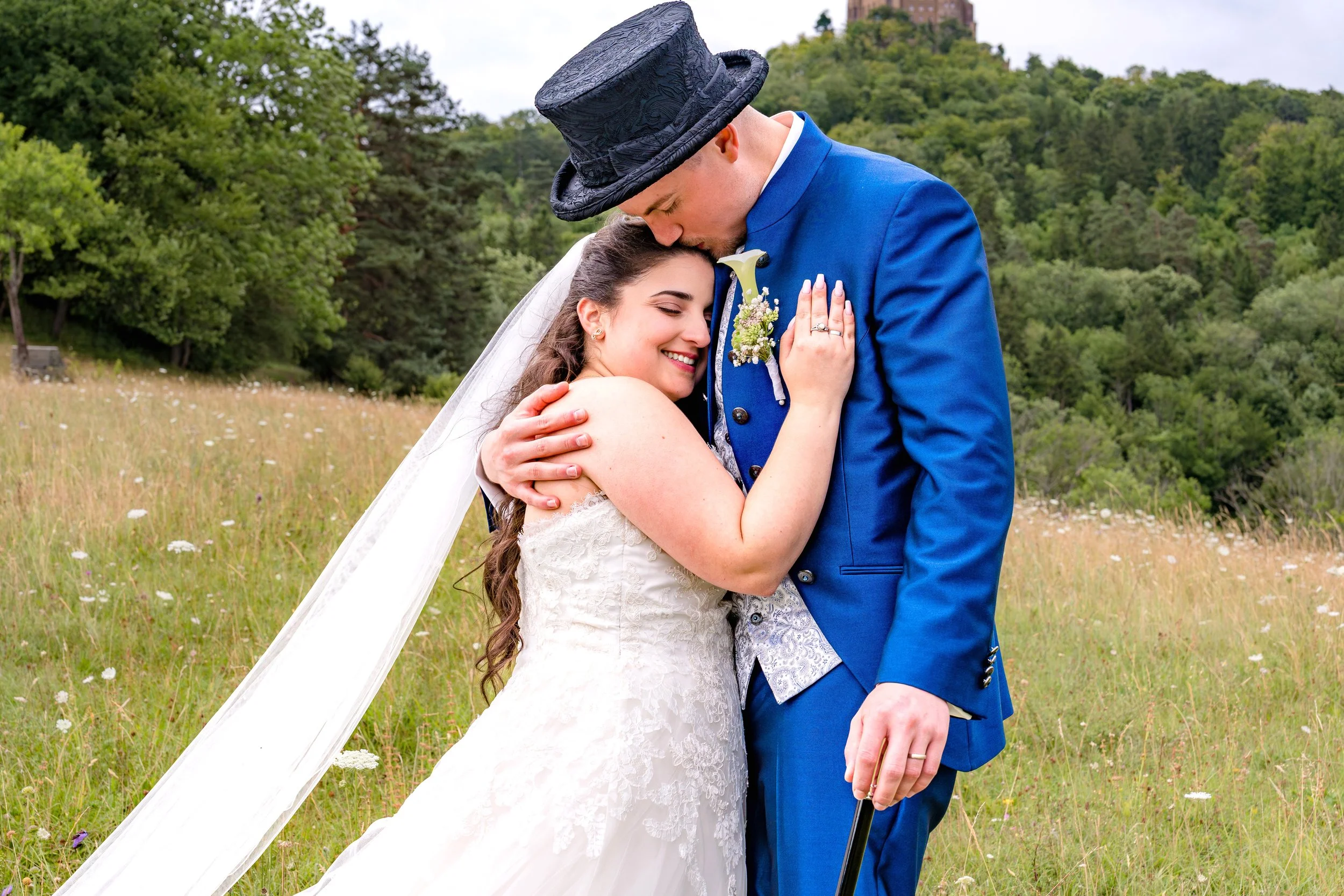 Ein Brautpaar umarmt sich im Grünen, die Braut trägt ein weißes Hochzeitskleid und der Bräutigam einen blauen Anzug mit Zylinder.