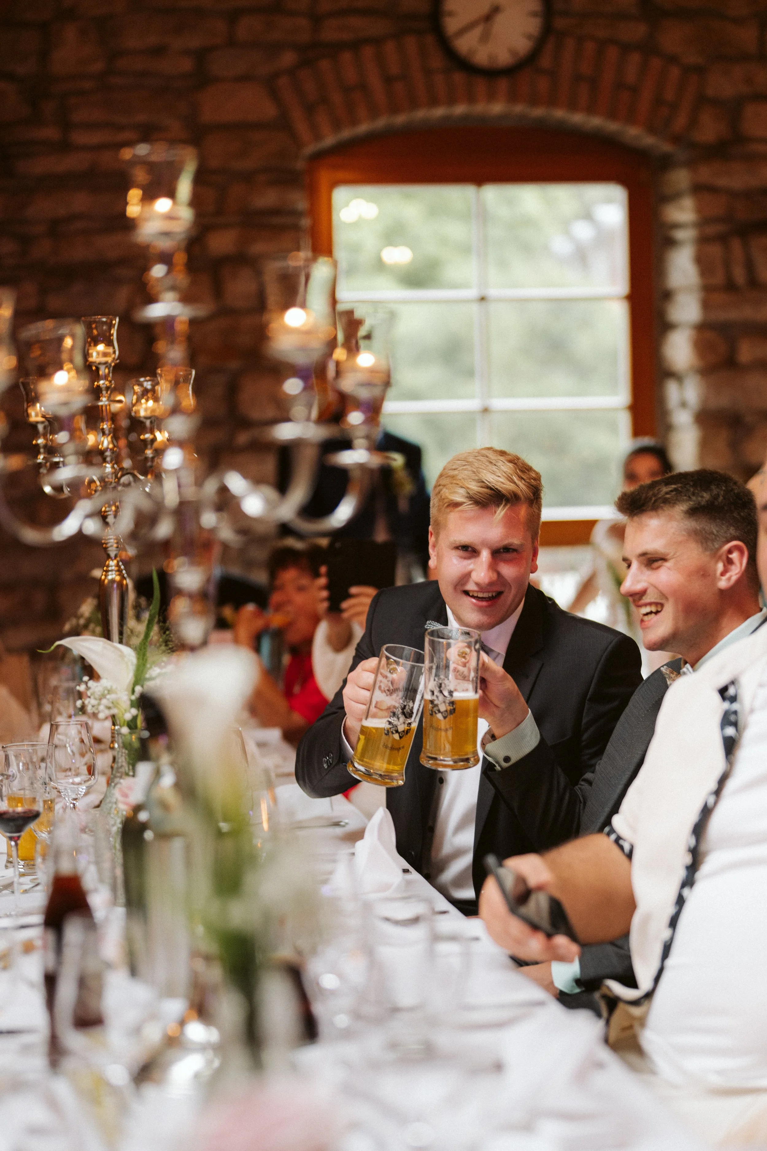 Junge Männer in Anzügen stoßen mit Bierkrügen an und lachen bei einem Festmahl in einem rustikalen Saal mit Ziegelwänden und Kerzen. Es sind mehrere Gäste zu sehen.