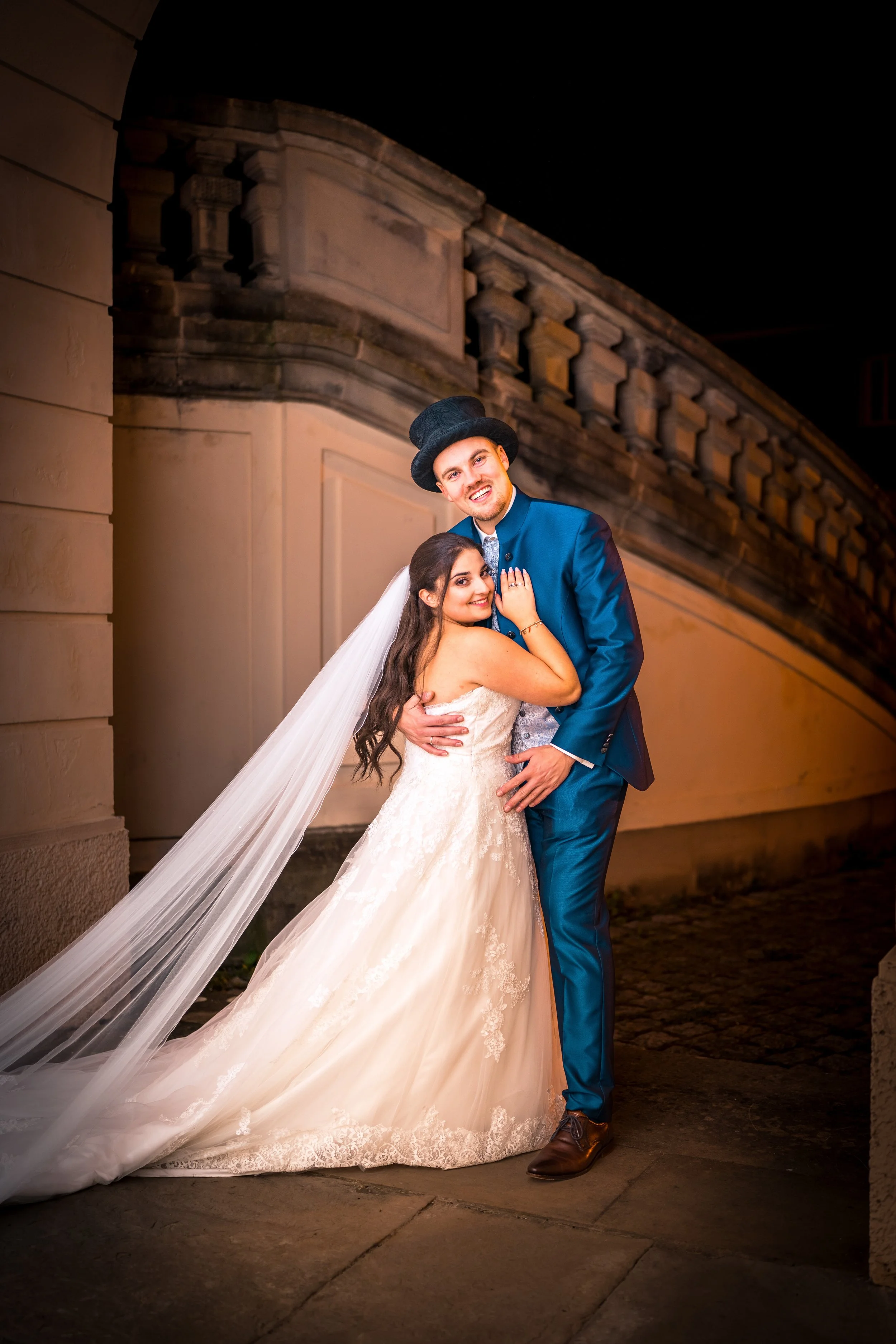 Ein glückliches Brautpaar posiert bei Nacht vor einer alten Steinmauer. Die Braut trägt ein weißes Hochzeitskleid mit Schleier, der Bräutigam trägt einen blauen Anzug und einen schwarzen Hut.