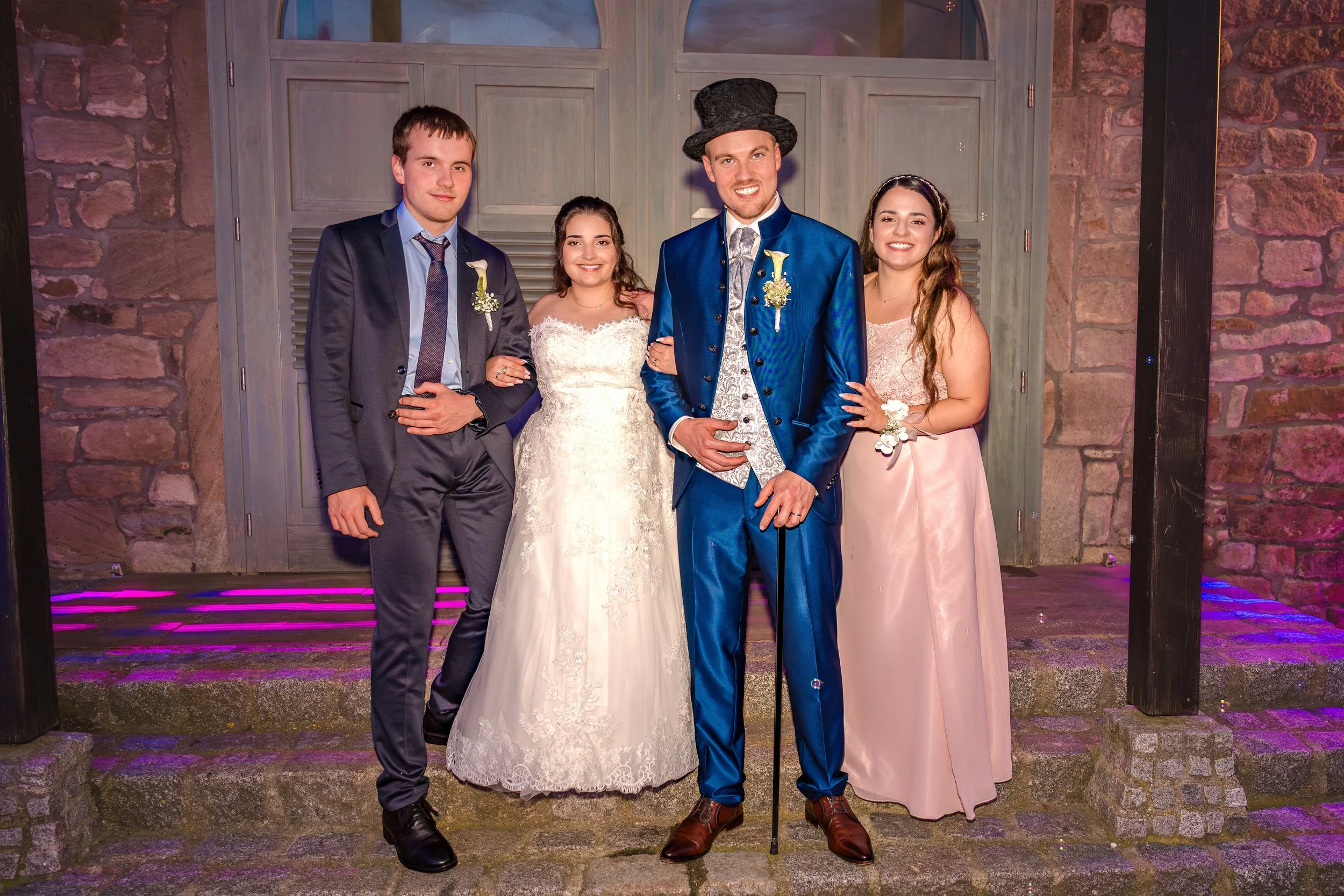 Hochzeitsgesellschaft bestehend aus vier Personen, zwei Männer und zwei Frauen, bei einer Feier im Freien vor einem Holzgebäude, mit Dekorationen und bunten Lichtern.