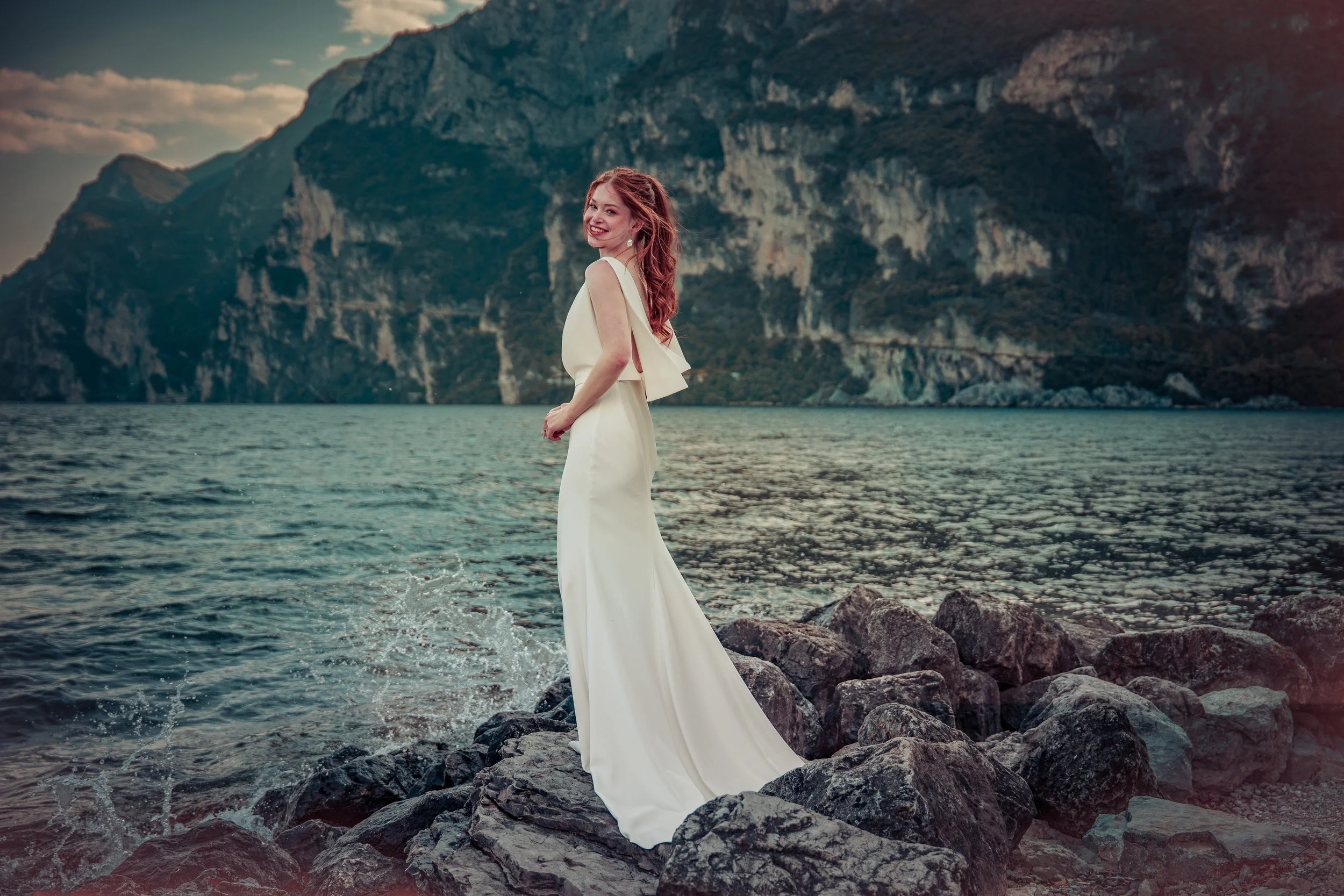 Frau in einem weißen Kleid steht auf Felsen am See, im Hintergrund Berge, bei Sonnenuntergang