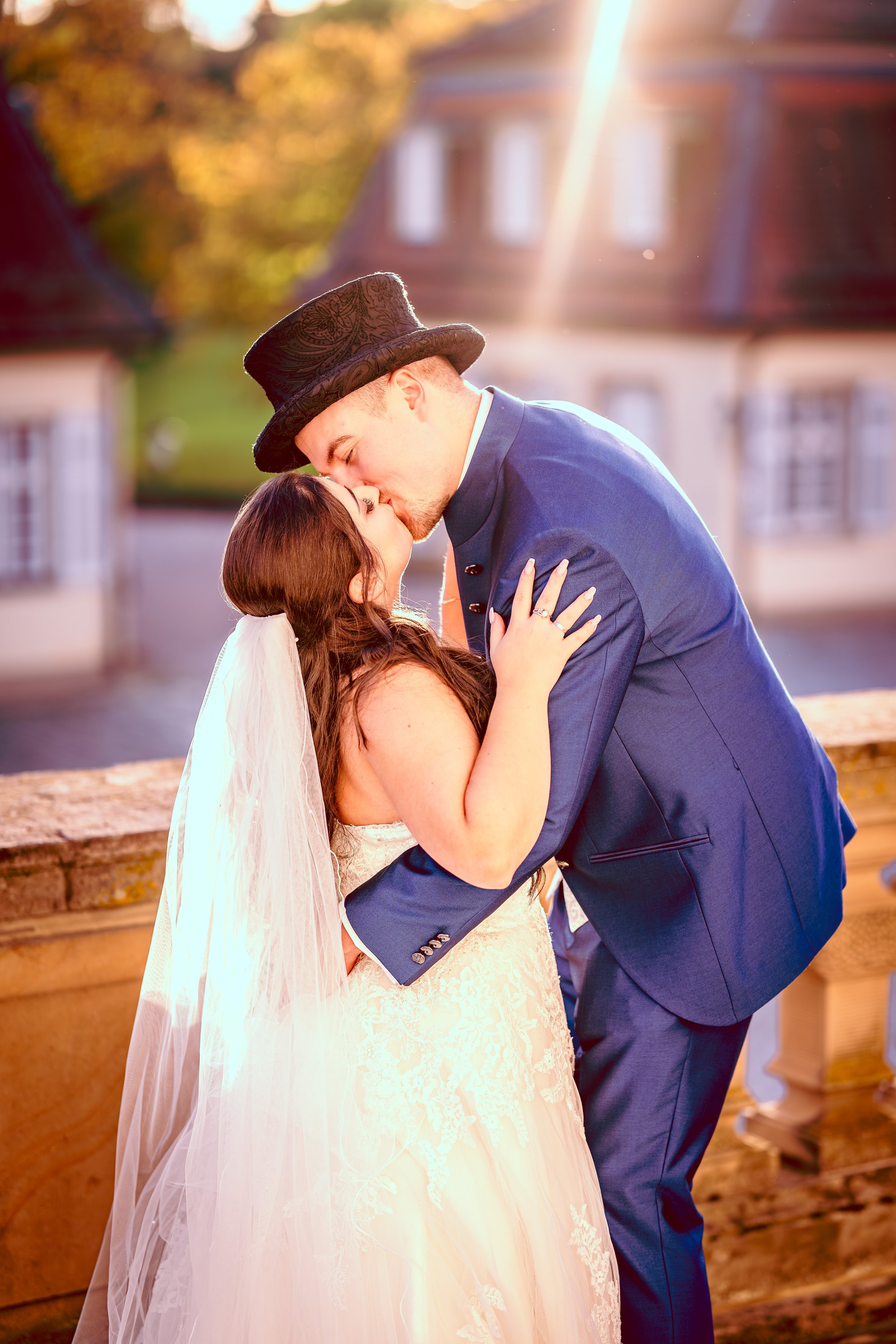 Ein frisch vermähltes Paar küsst sich bei Sonnenuntergang auf einer Brücke, die Braut trägt ein weißes Hochzeitskleid und Schleier, der Bräutigam trägt einen blauen Anzug mit Zylinder.