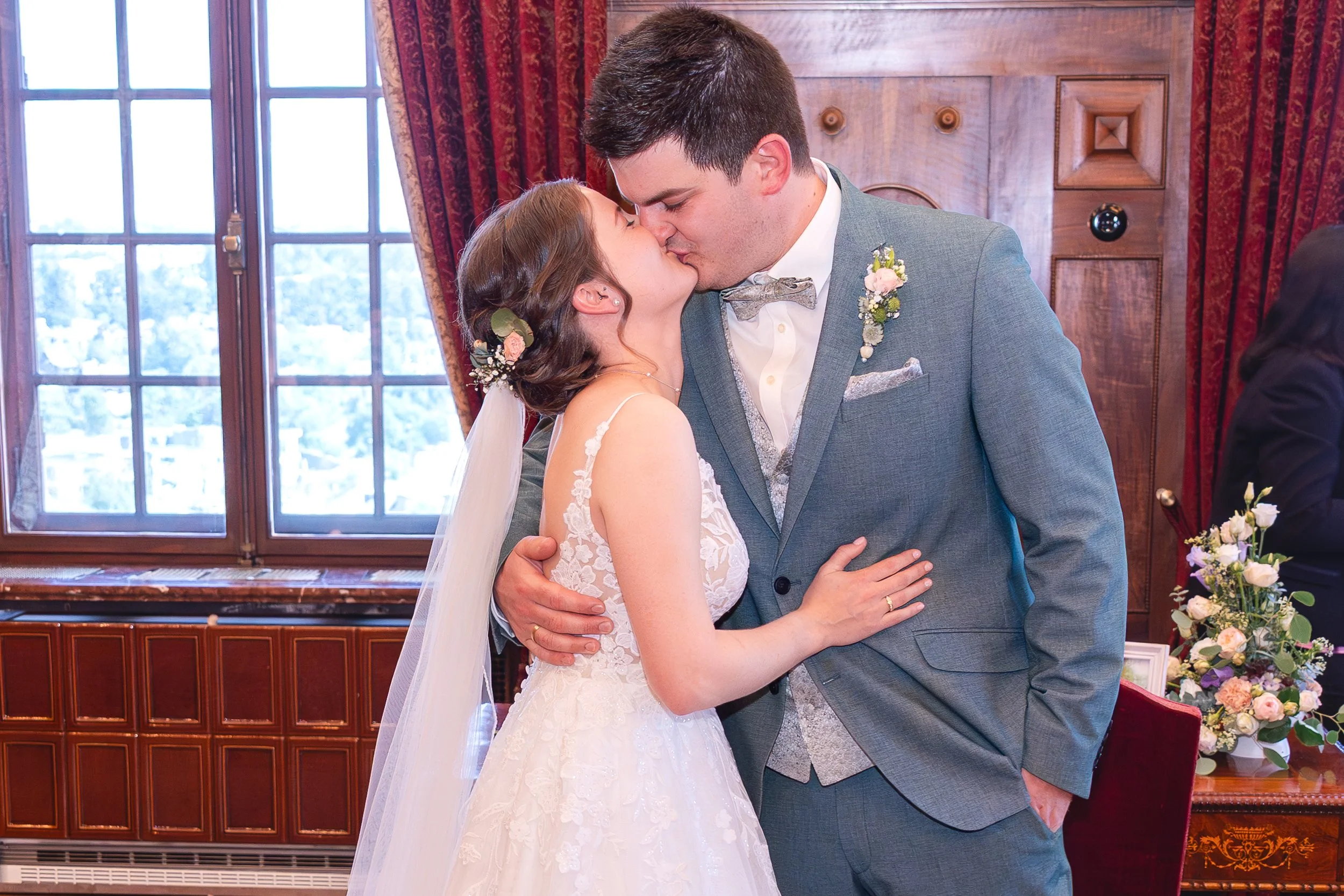 Junges Brautpaar während der Hochzeitszeremonie umarmt sich und küsst sich in einem festlichen Raum mit Holzpaneele und großen Fenstern.