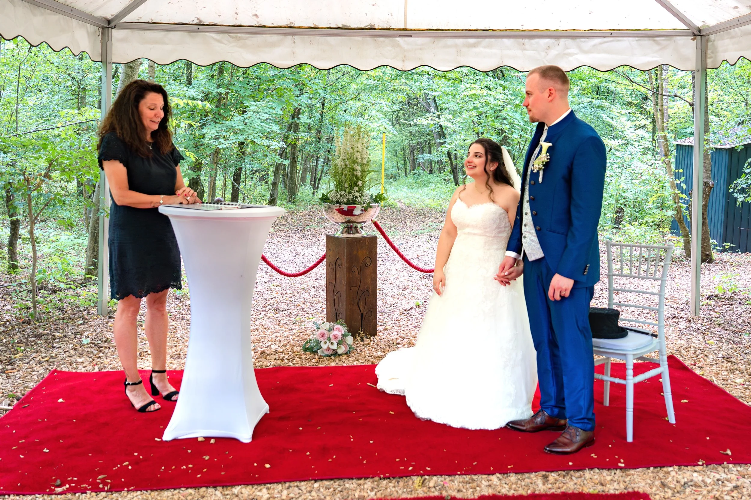 Hochzeit im Freien unter einem Zelt, Braut und Bräutigam halten Händchen während der Trauung, eine Frau liest aus einem Buch, rote Samtband vor einem Blumenarrangement, roter Teppich auf dem Boden.
