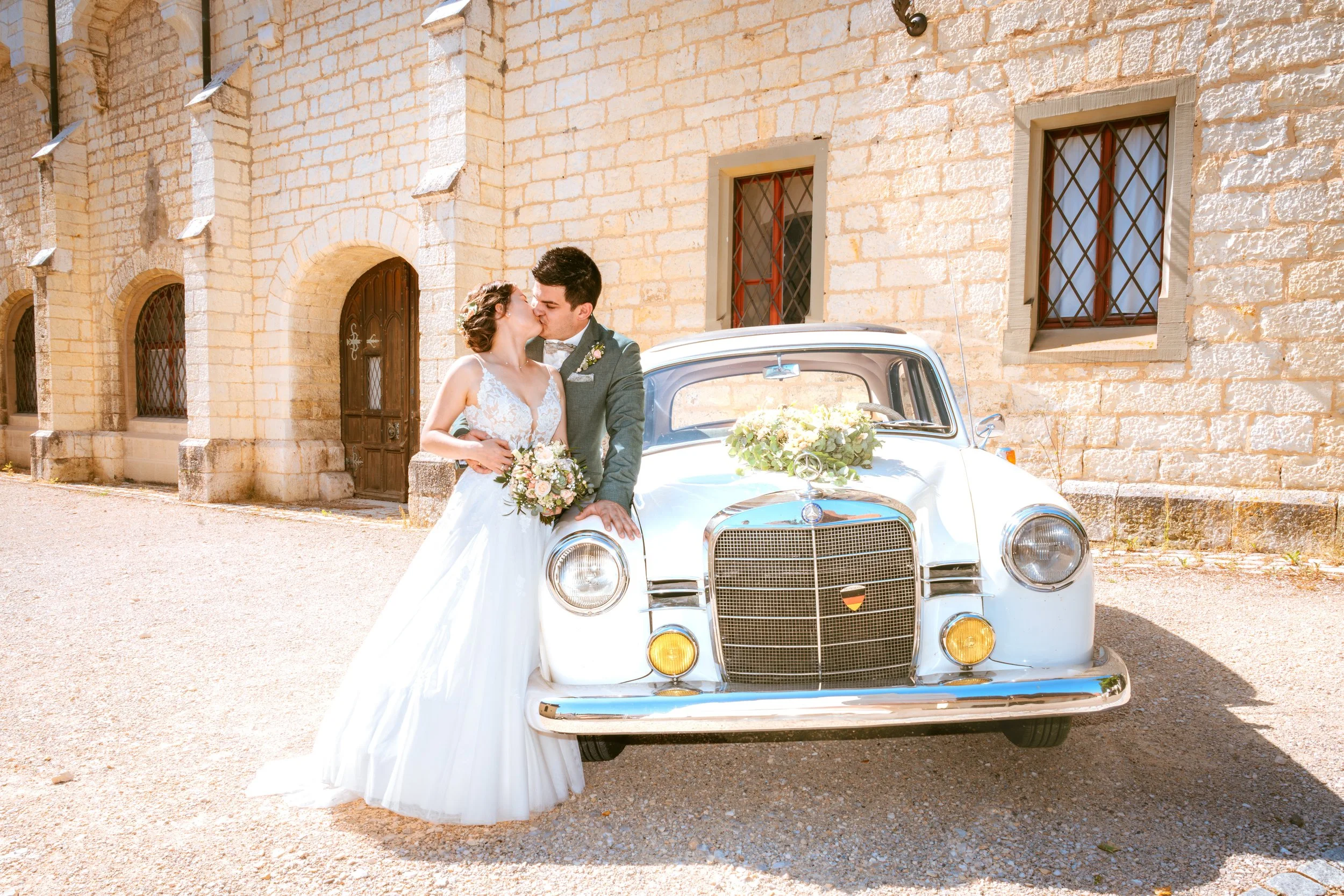 Ein Brautpaar bei ihrer Hochzeit, das sich liebevoll küsst, steht neben einem klassischen weißen Auto mit Blumenverzierung vor einem Steinmauern-Hintergrund.