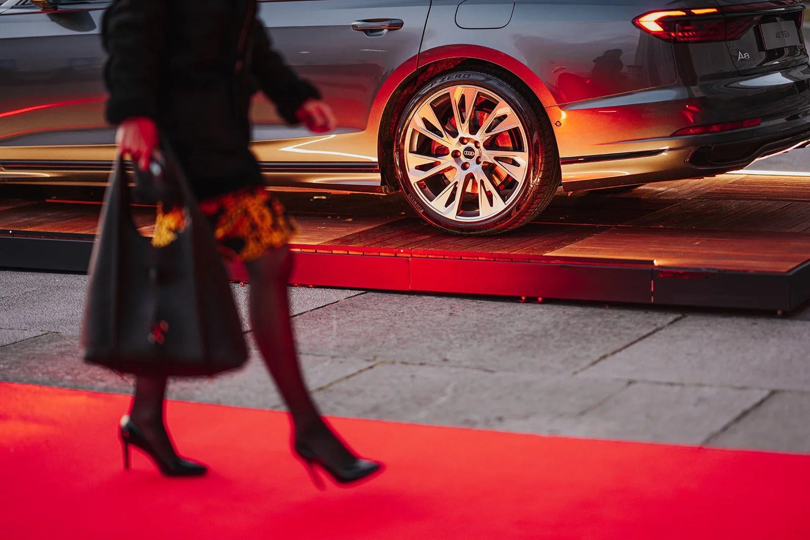 A car on display at an auto show, with a woman wearing high heels walking past in front of the red carpet.