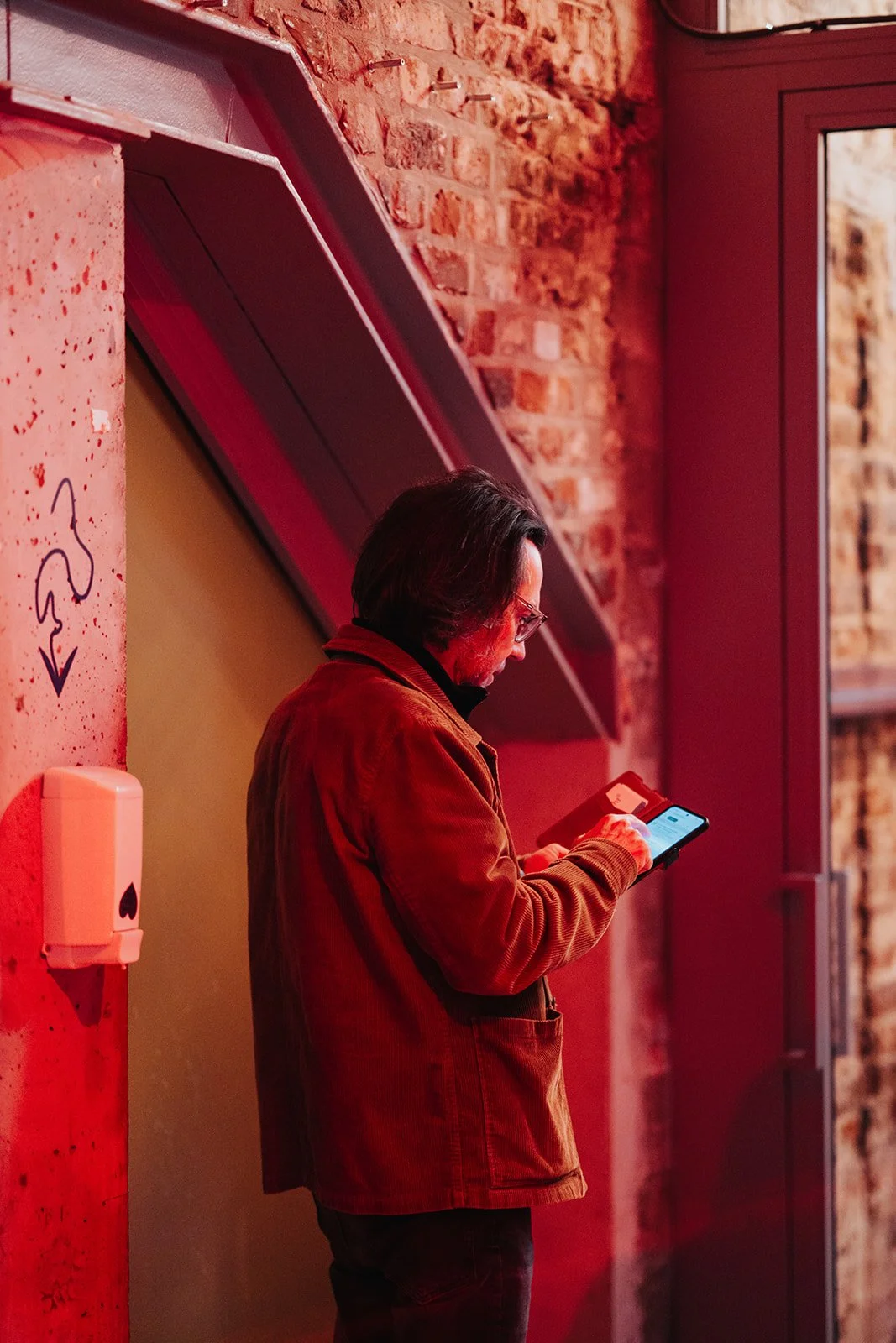 A man with glasses and dark hair, wearing a brown jacket, stands against a brick wall in a dimly lit room with red lighting, focused on his phone.