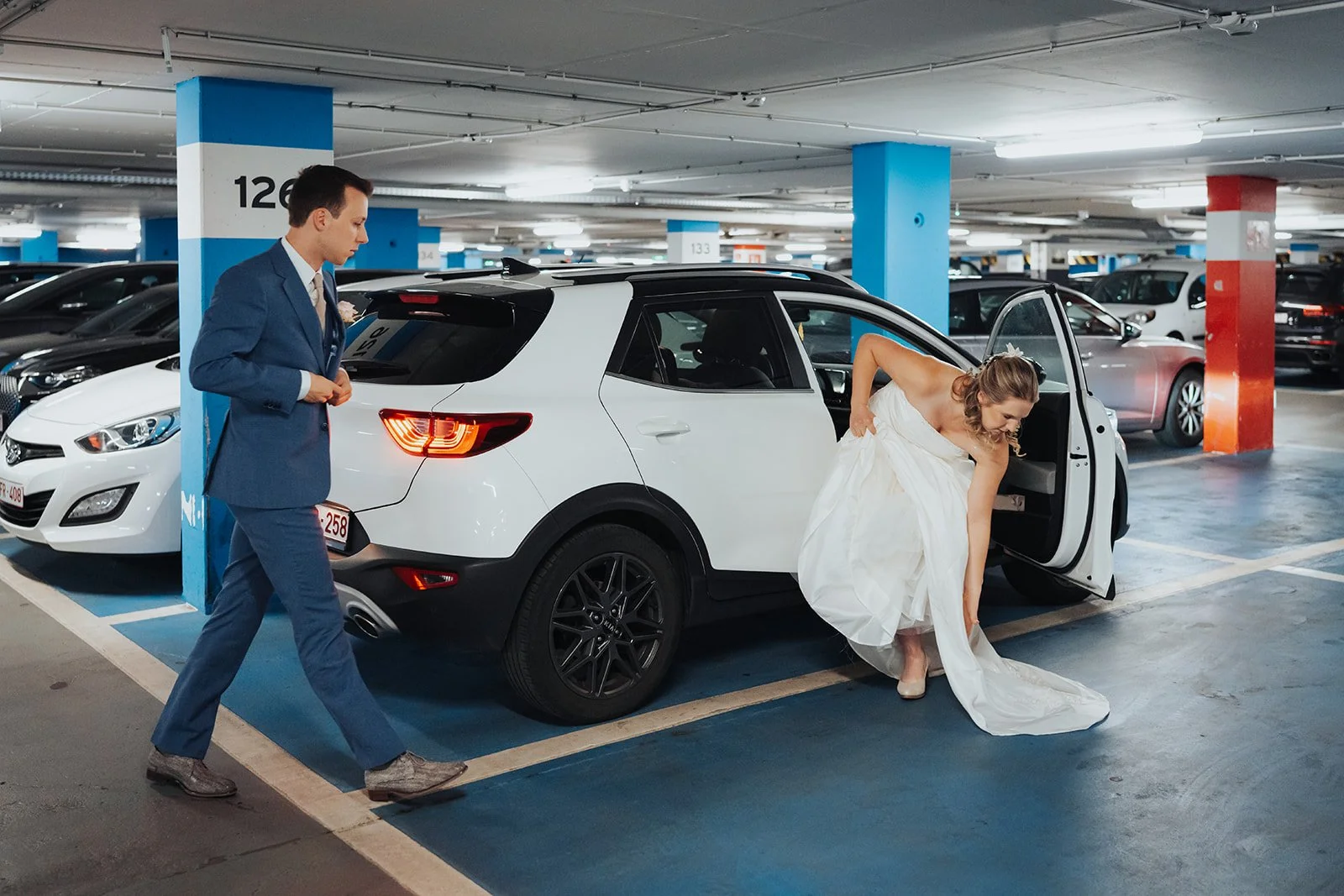 A bride in a white wedding dress is stepping out of a white SUV in an indoor parking garage, as the groom in a blue suit walks nearby, buttoning his jacket.