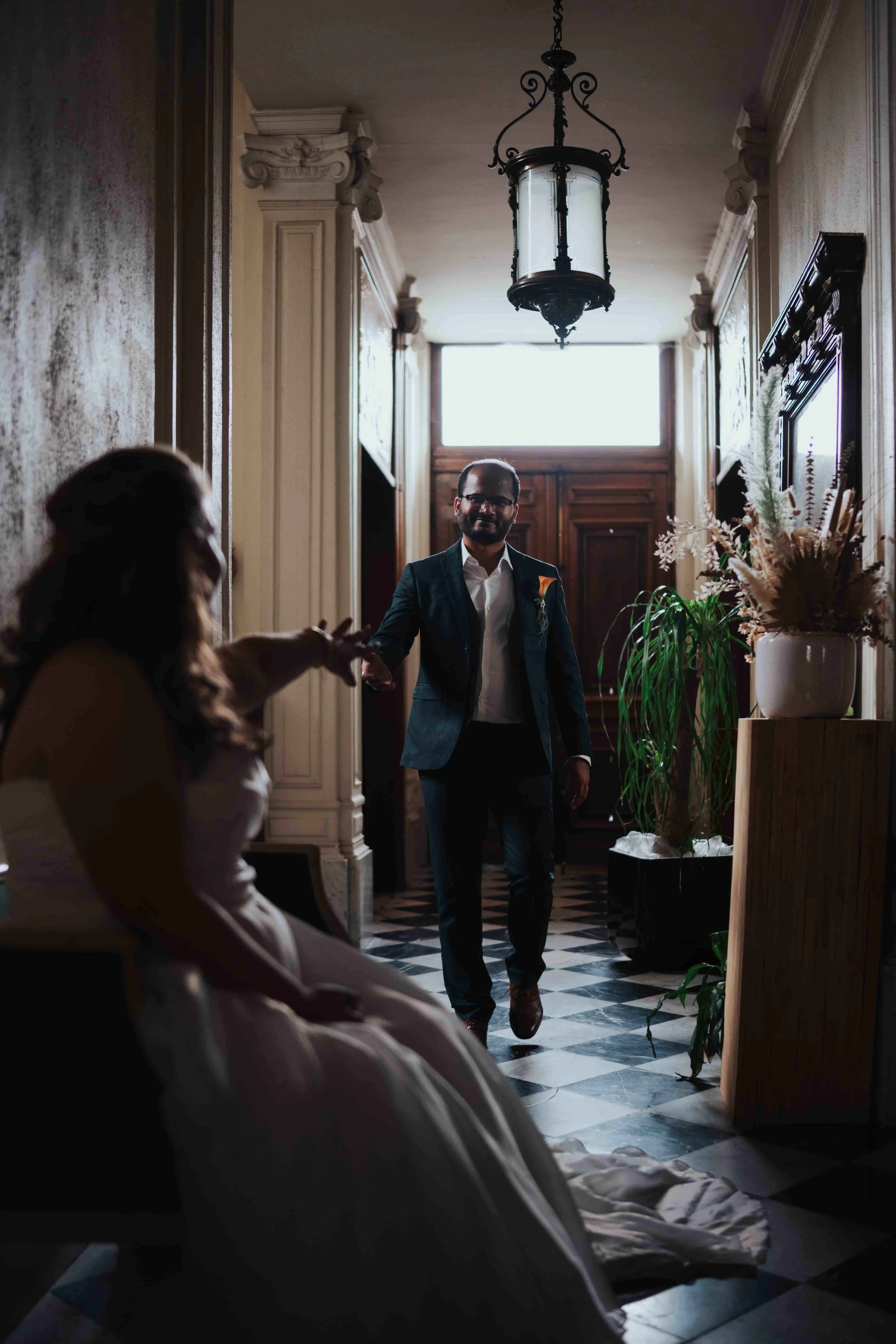 A man in a suit walking into a room where a woman in a wedding dress is sitting. The room features ornate woodwork, plants, and vintage lighting.