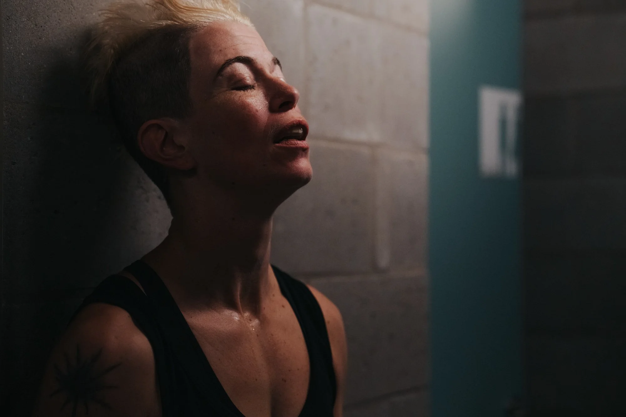 A woman with short blonde hair leaning against a concrete wall, appearing exhausted or emotional, with her eyes closed and sweat on her face and neck.