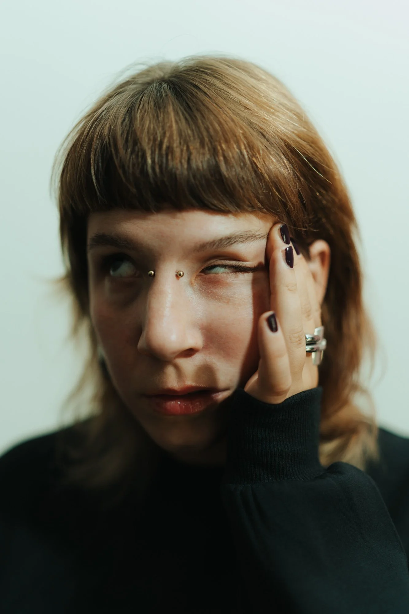 A person with light brown hair and multiple facial piercings, including two small studs at the nose bridge, making a face with one eye closed and the other eye rolled up, with their hand covering part of their face.