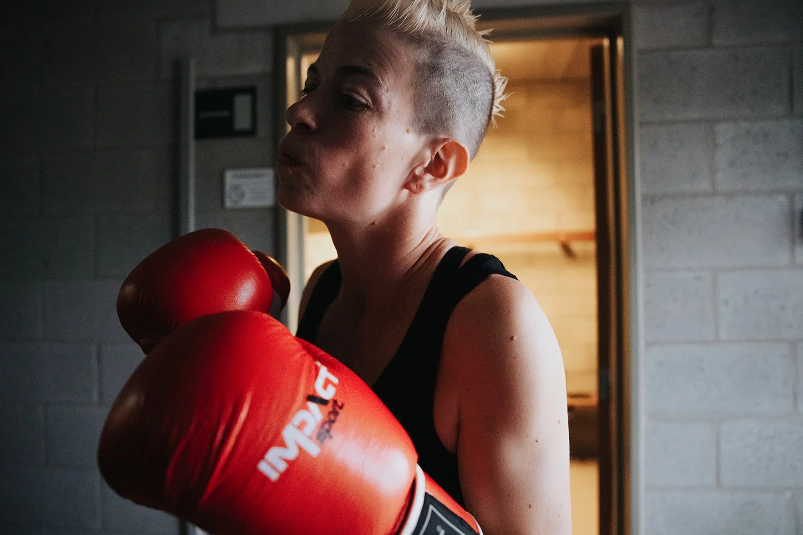 A female boxer with short blonde hair and a black sports top, wearing red boxing gloves, standing in a gym.