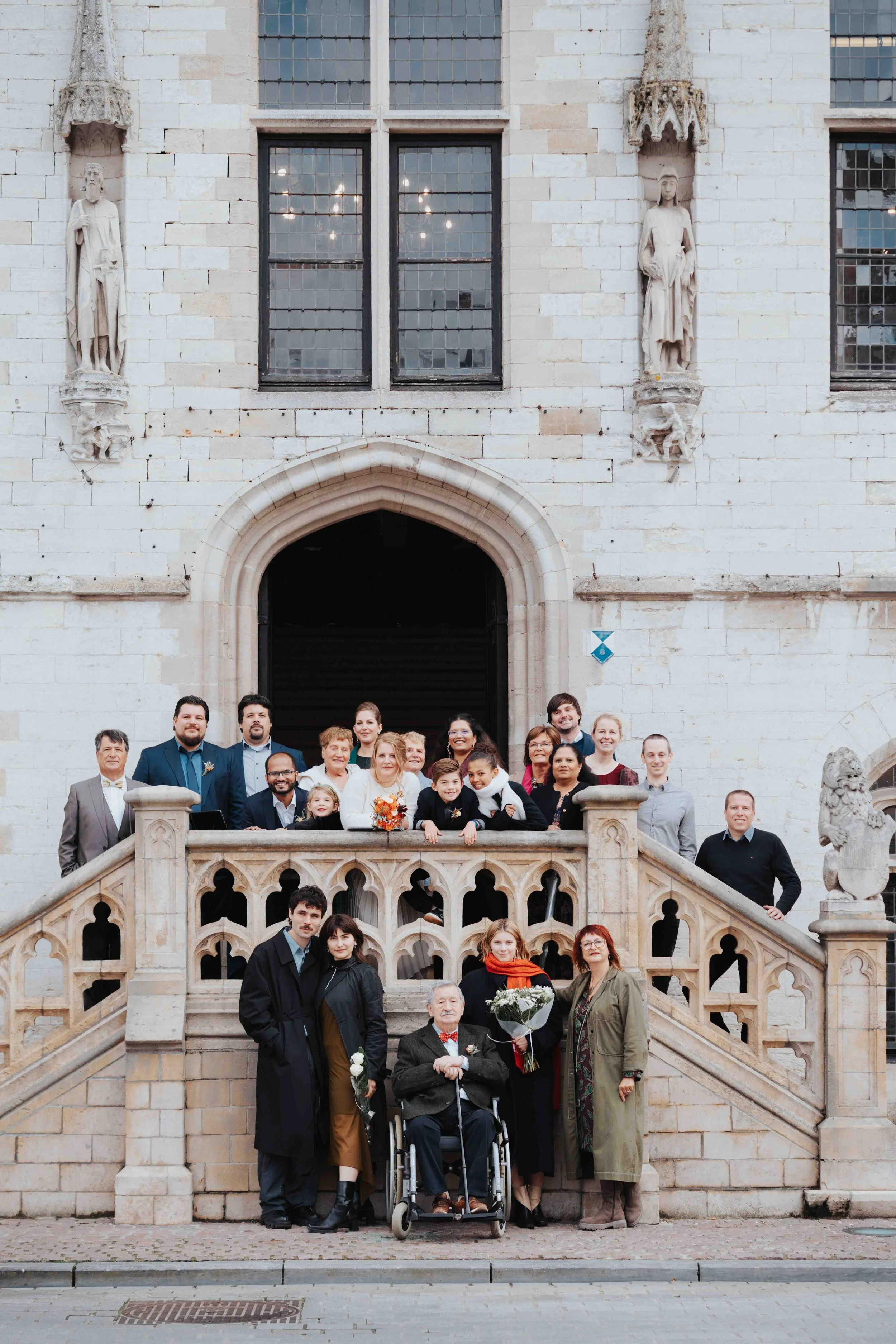 A large group of people, including children, adults, and elderly, gathered on the steps of a historic stone building with statues and large stained-glass windows, for a group photo during a wedding.