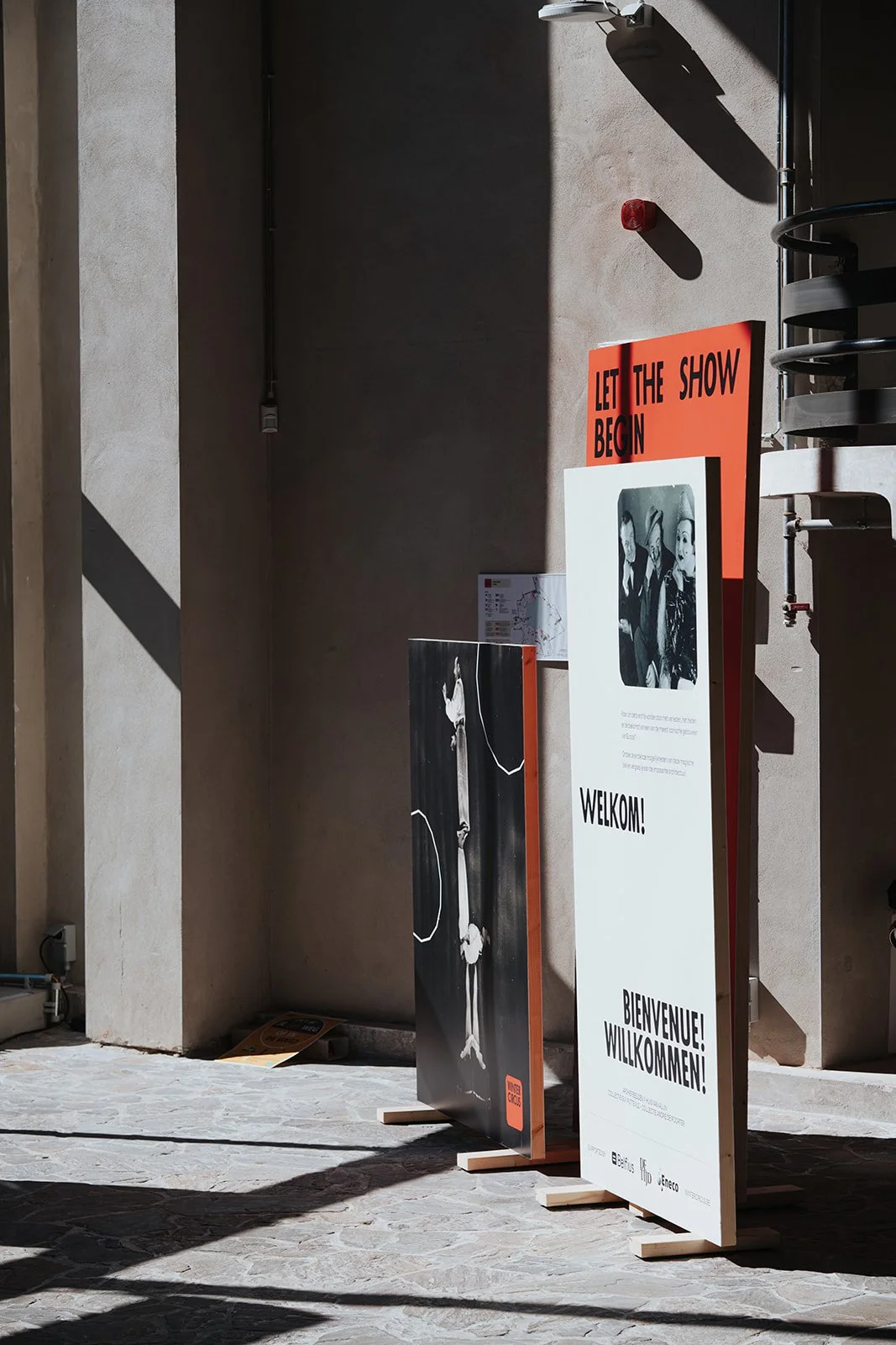 Indoor area with signs, including one reading "LET THE SHOW BEGIN" and welcoming visitors in multiple languages, with shadow patterns on the floor and wall.