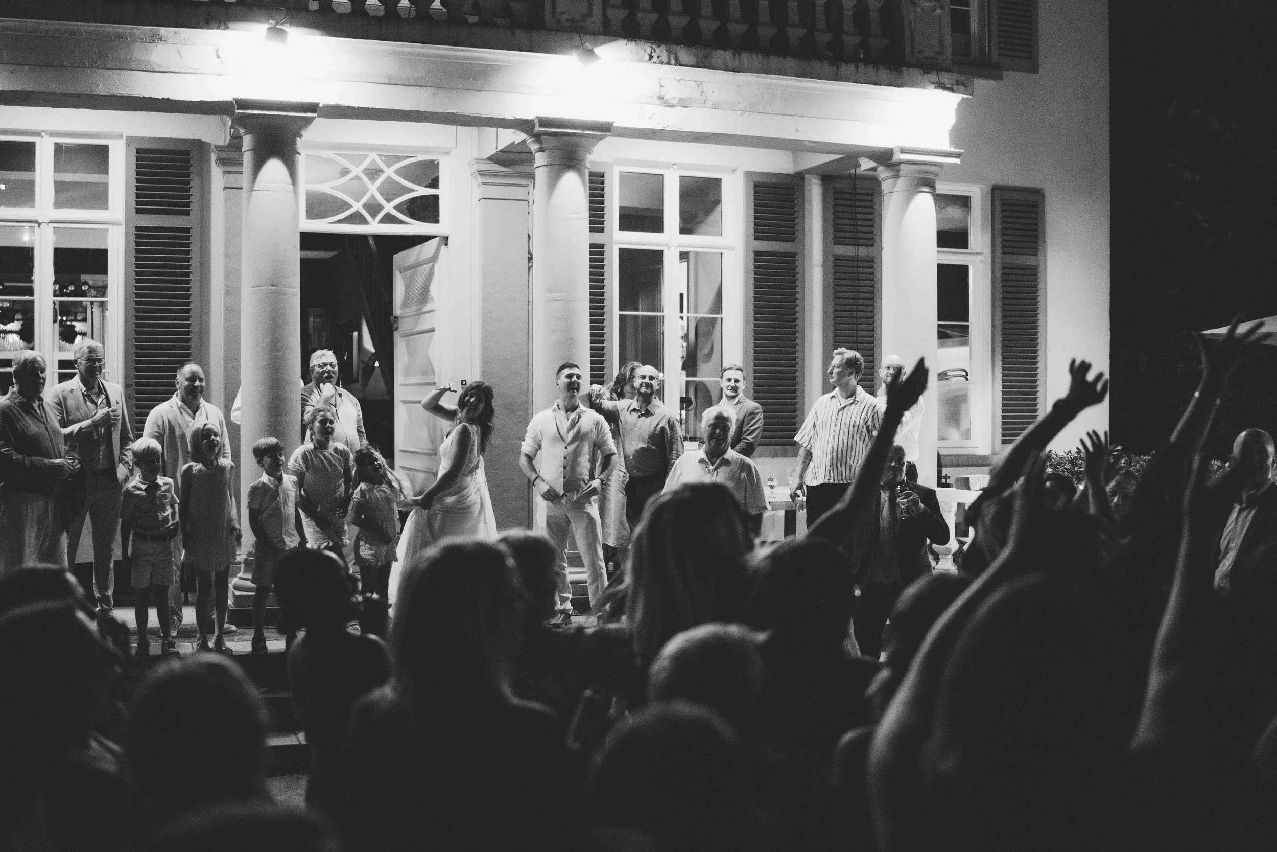 Black and white photo of a group of people gathered outside a building at night, the bride is throwing her flowers, with a few people in the audience raising their hands to catch a boquet at a wedding reception.