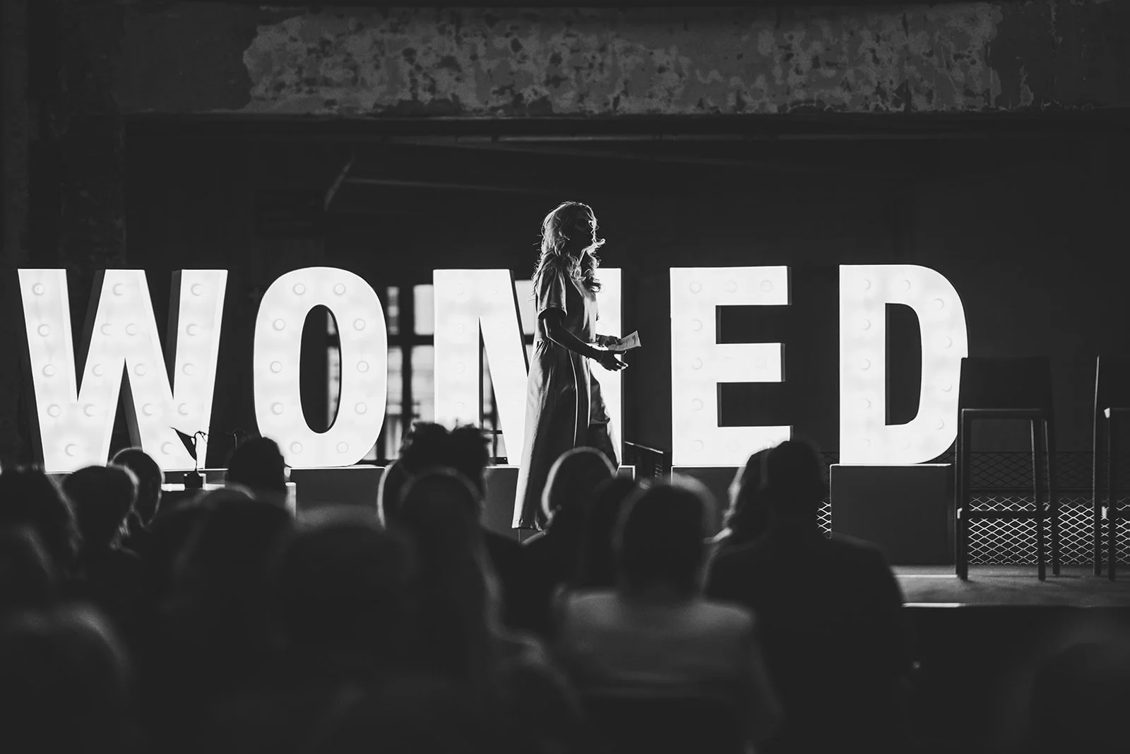 A woman speaking on stage in front of large illuminated letters spelling "WONED" with an audience watching.
