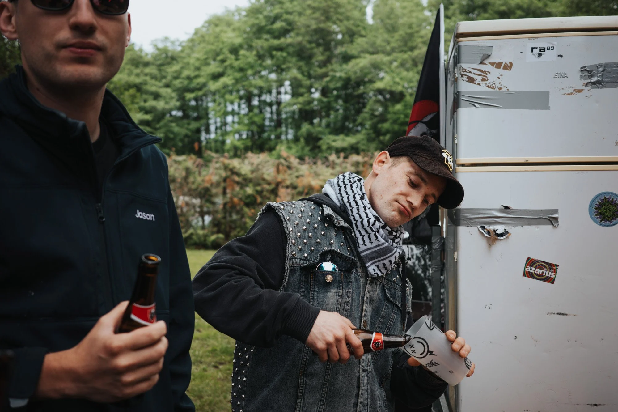 Young man wearing a black cap and a studded denim vest pouring a drink from a bottle into a paper cup, with another person holding a beer bottle in the foreground. They are outdoors near a white trailer with stickers, and a flag is visible behind the