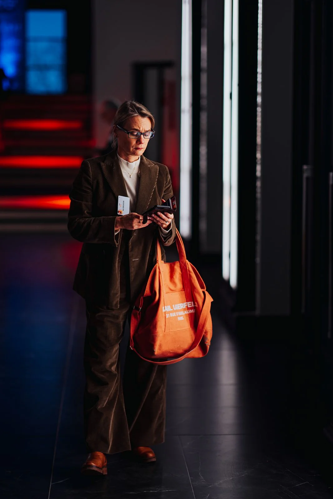 A woman in a brown suit and glasses is standing in an indoor space with dark flooring and illuminated vertical light panels. She is looking at her phone, holding a credit card, and carrying an orange Karl Lagerfeld tote bag.