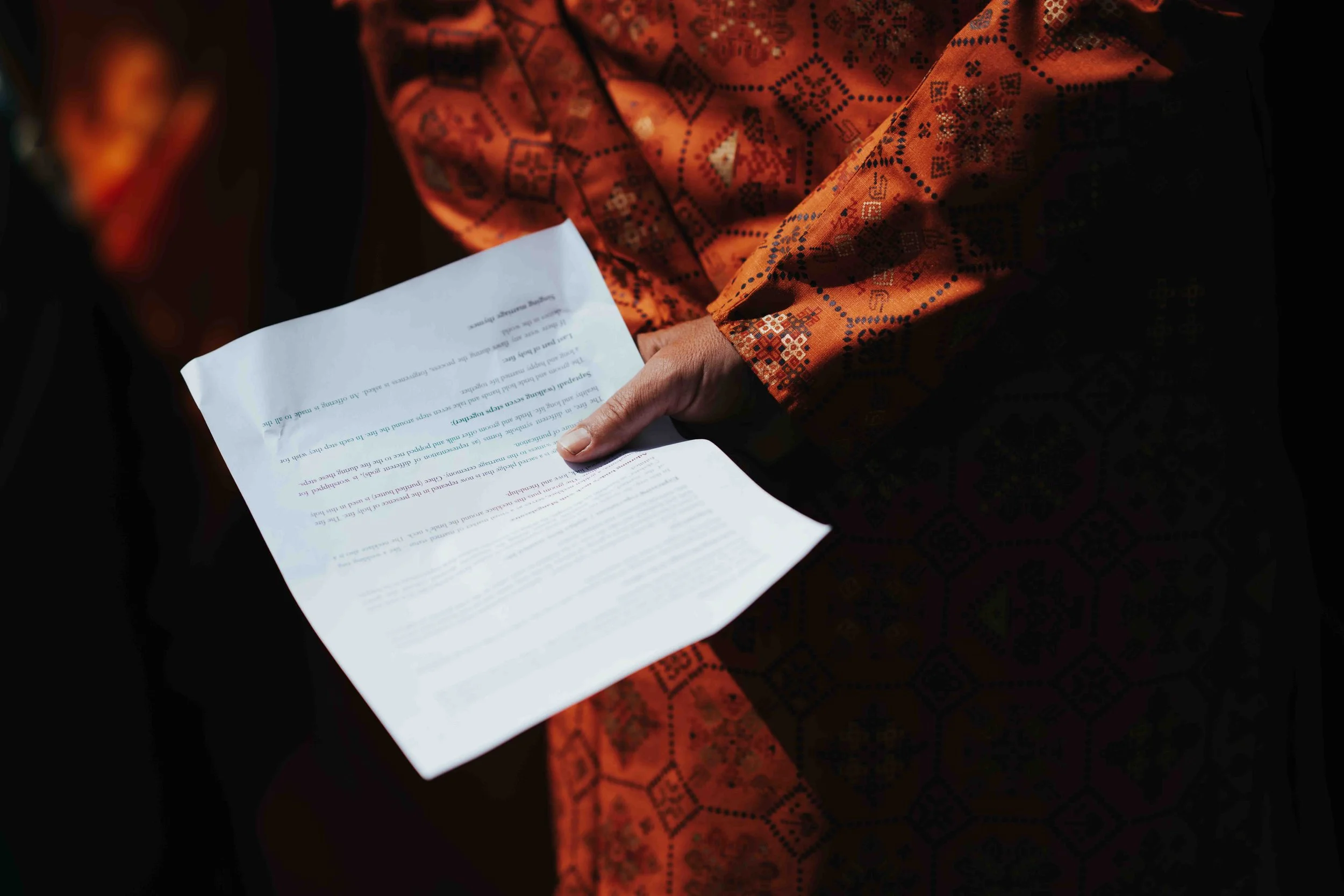 Person wearing an orange, patterned traditional indian garment holding a piece of paper with printed text.