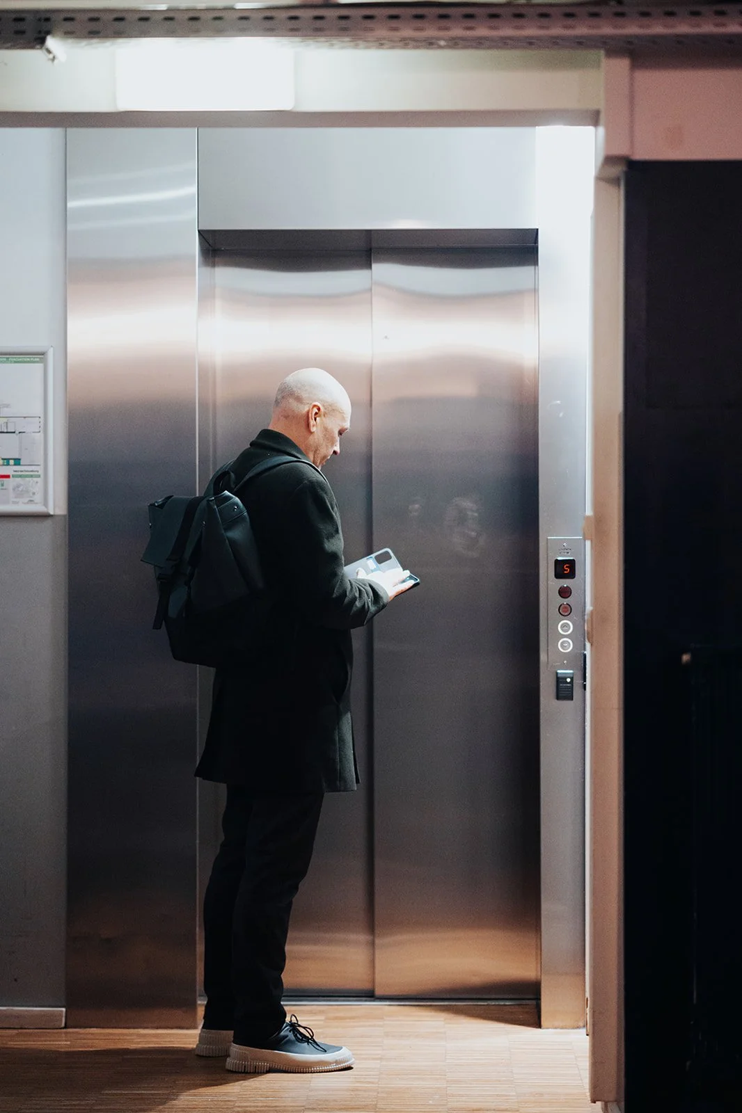 A bald man in a black coat and black pants with a backpack waiting in front of an elevator door, looking at his phone.