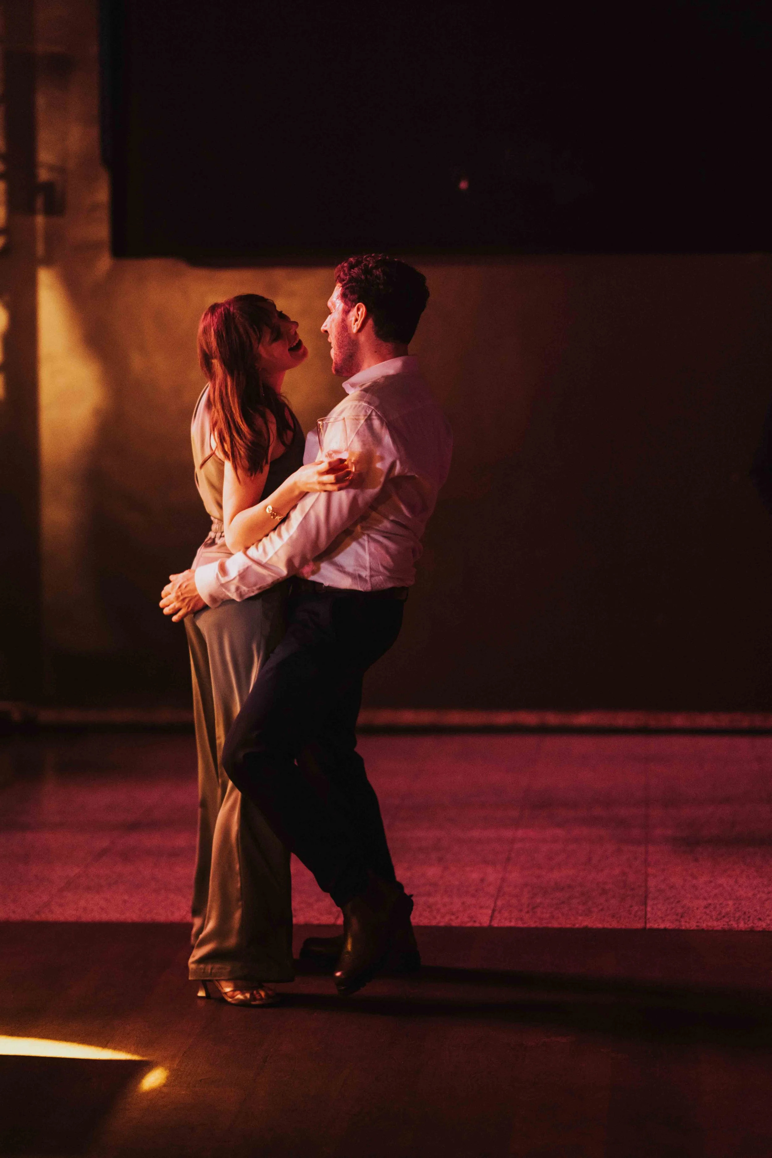 A man and woman are dancing closely at a dimly lit event, with the woman holding a glass of wine.