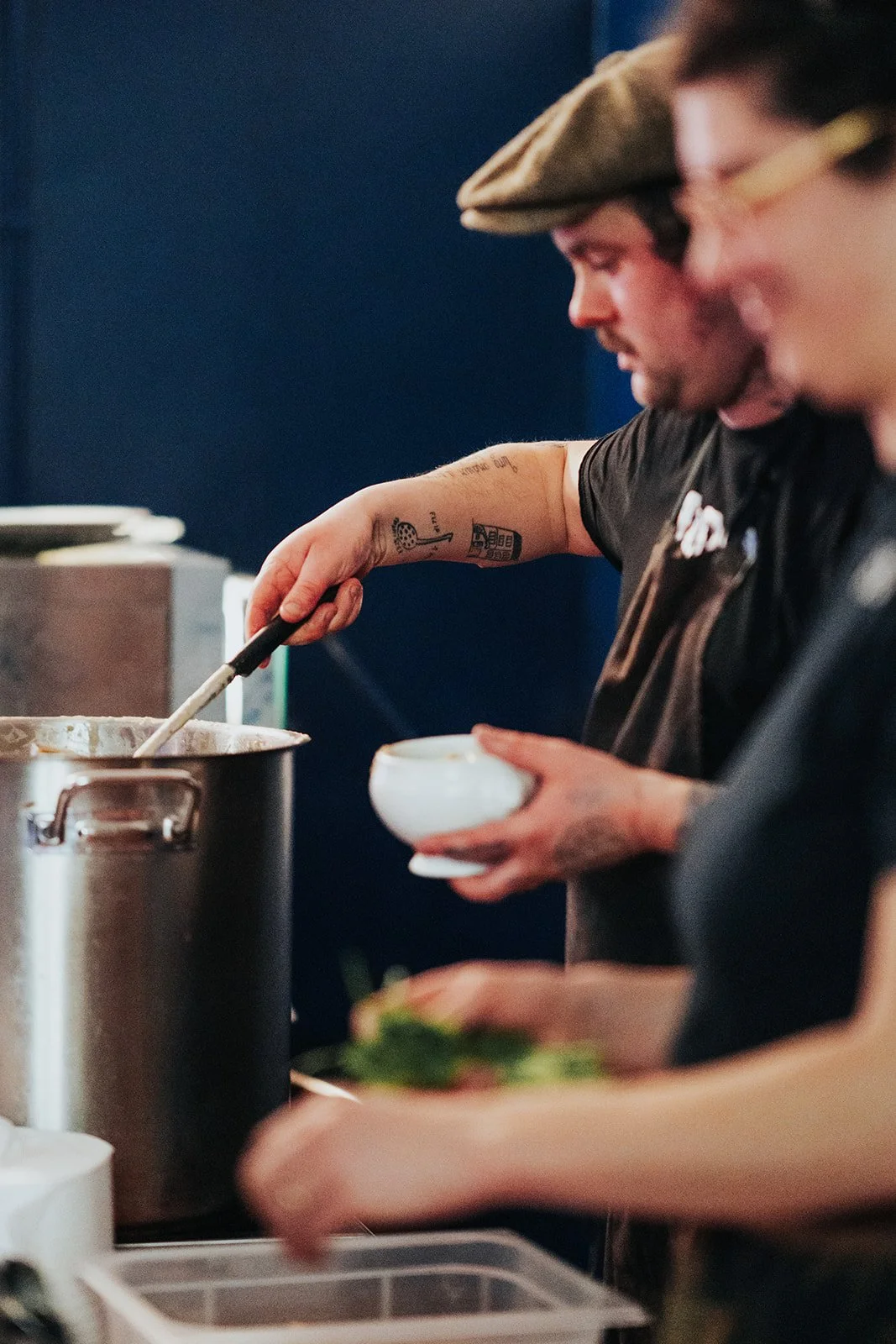 Two people preparing food in a kitchen, one stirring a pot and holding a bowl, the other chopping vegetables.